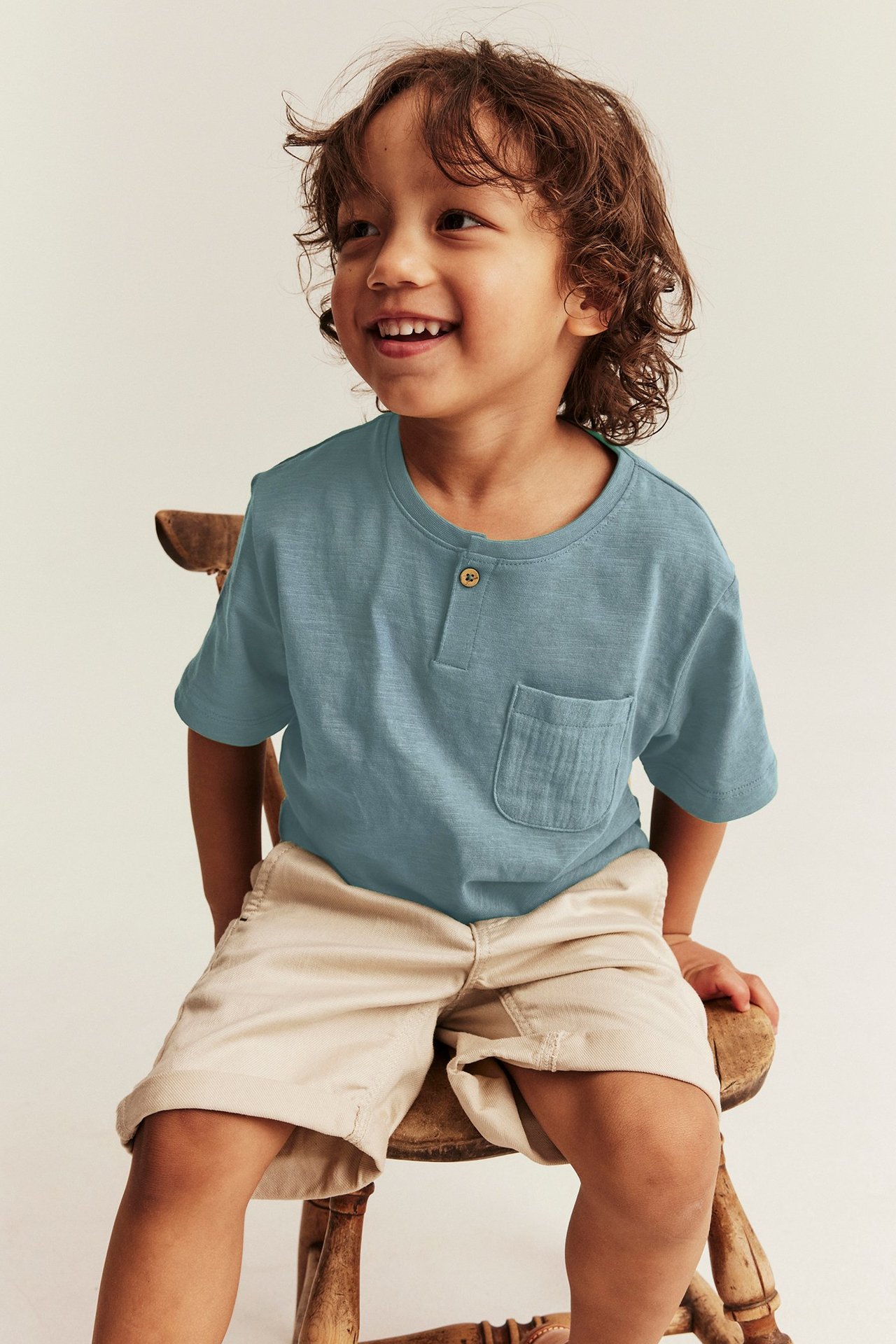 Seitenansicht: Ein Kind trägt ein dunkeltürkises Baumwoll-T-Shirt mit Knopf, Tasche und beigen Shorts.