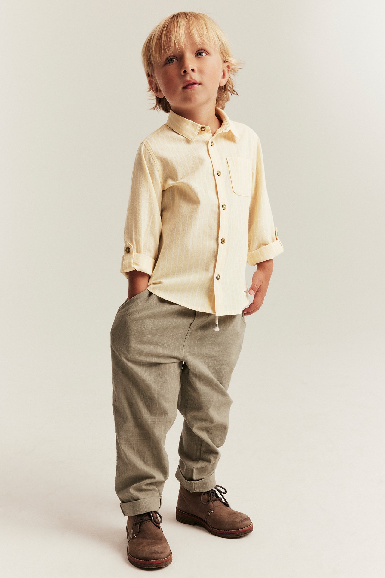 Front view of a kid in a light yellow striped cotton shirt with rolled-up sleeves and light brown trousers.
