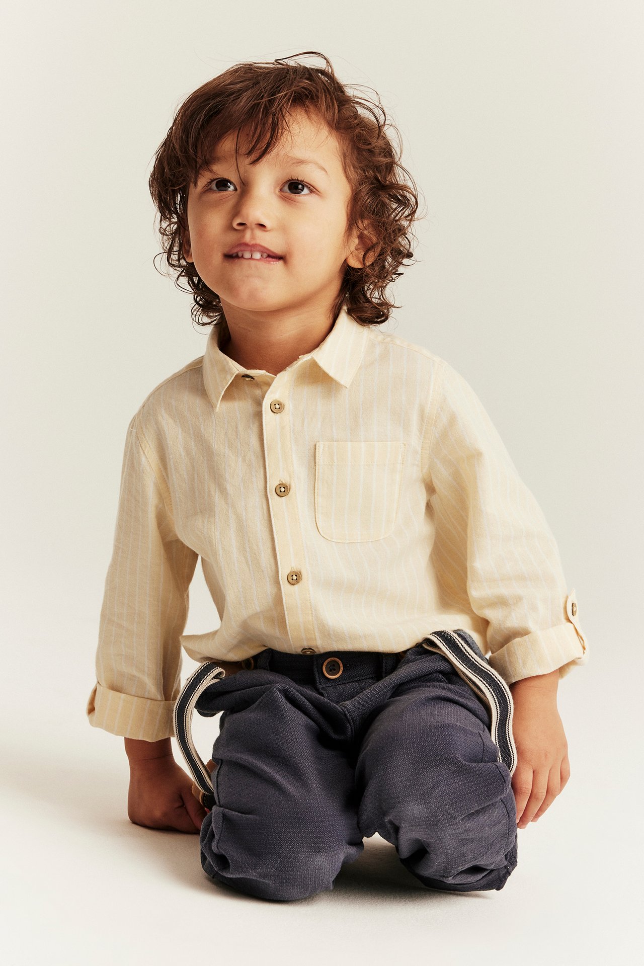 Front view of a kid wearing a light yellow striped cotton shirt with rolled-up sleeves and dark blue trousers.