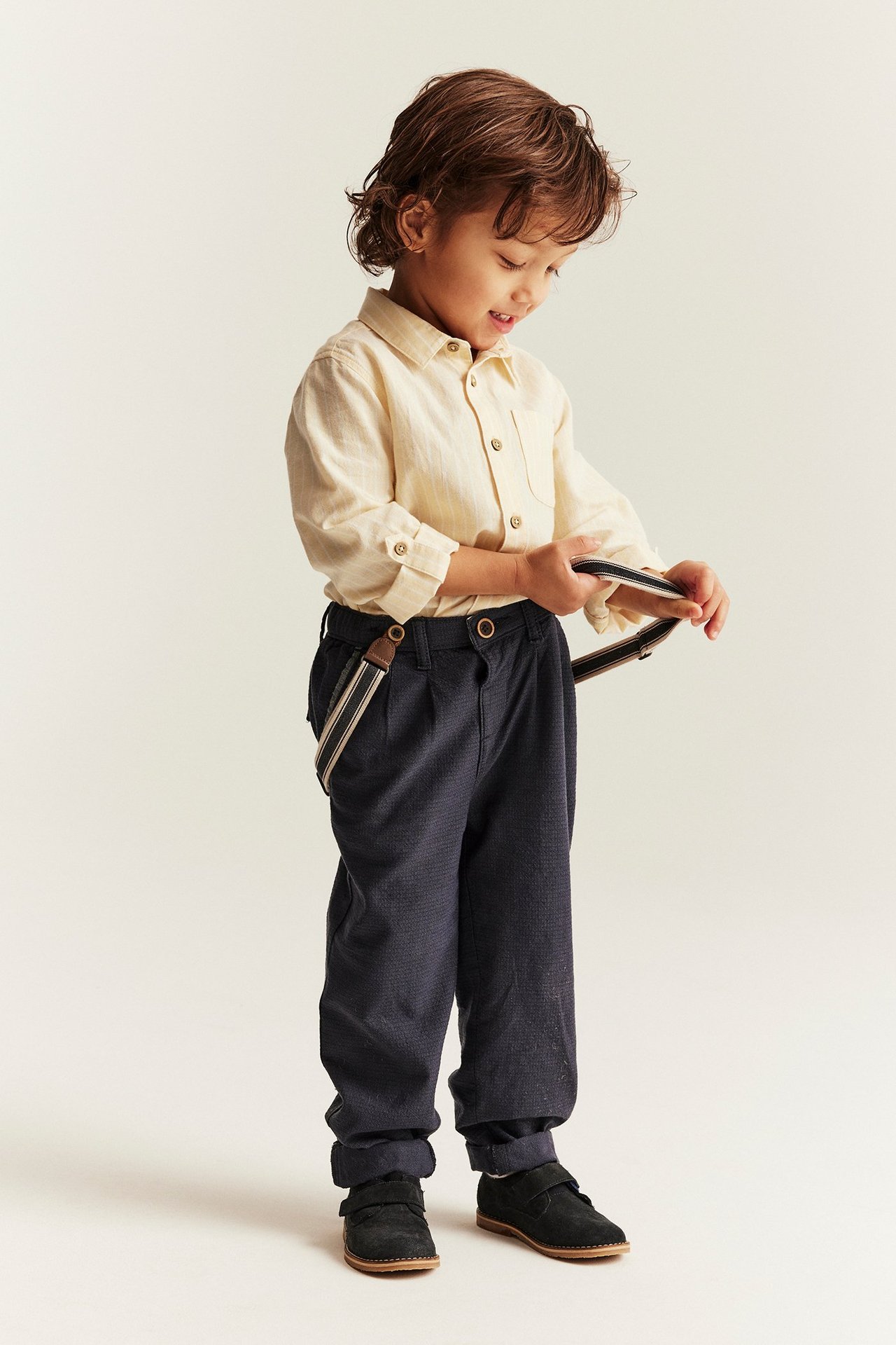 Front view: Kid wears a light yellow striped cotton shirt, dark blue trousers with suspenders, and black shoes.