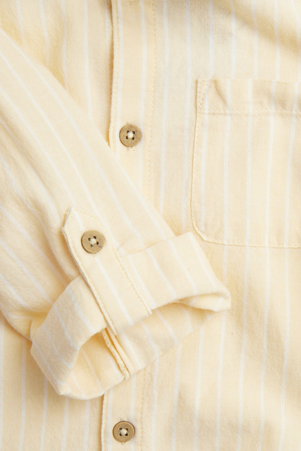 Close-up of a yellow striped woven cotton shirt with a rolled-up sleeve, front buttons, and chest pocket.