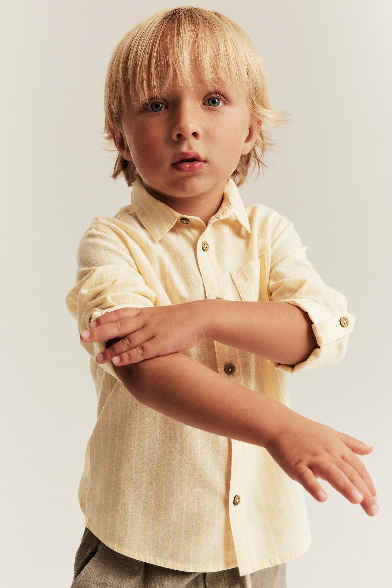 Front view of a kid wearing a light yellow striped woven cotton shirt with rolled-up sleeves and beige trousers.