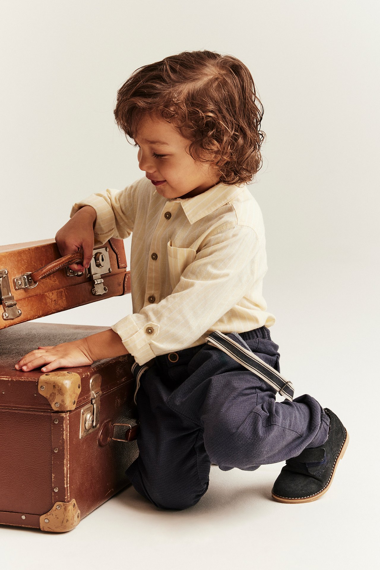 Front view: Kid in light yellow striped cotton shirt, dark pants, black shoes, opening brown suitcase.