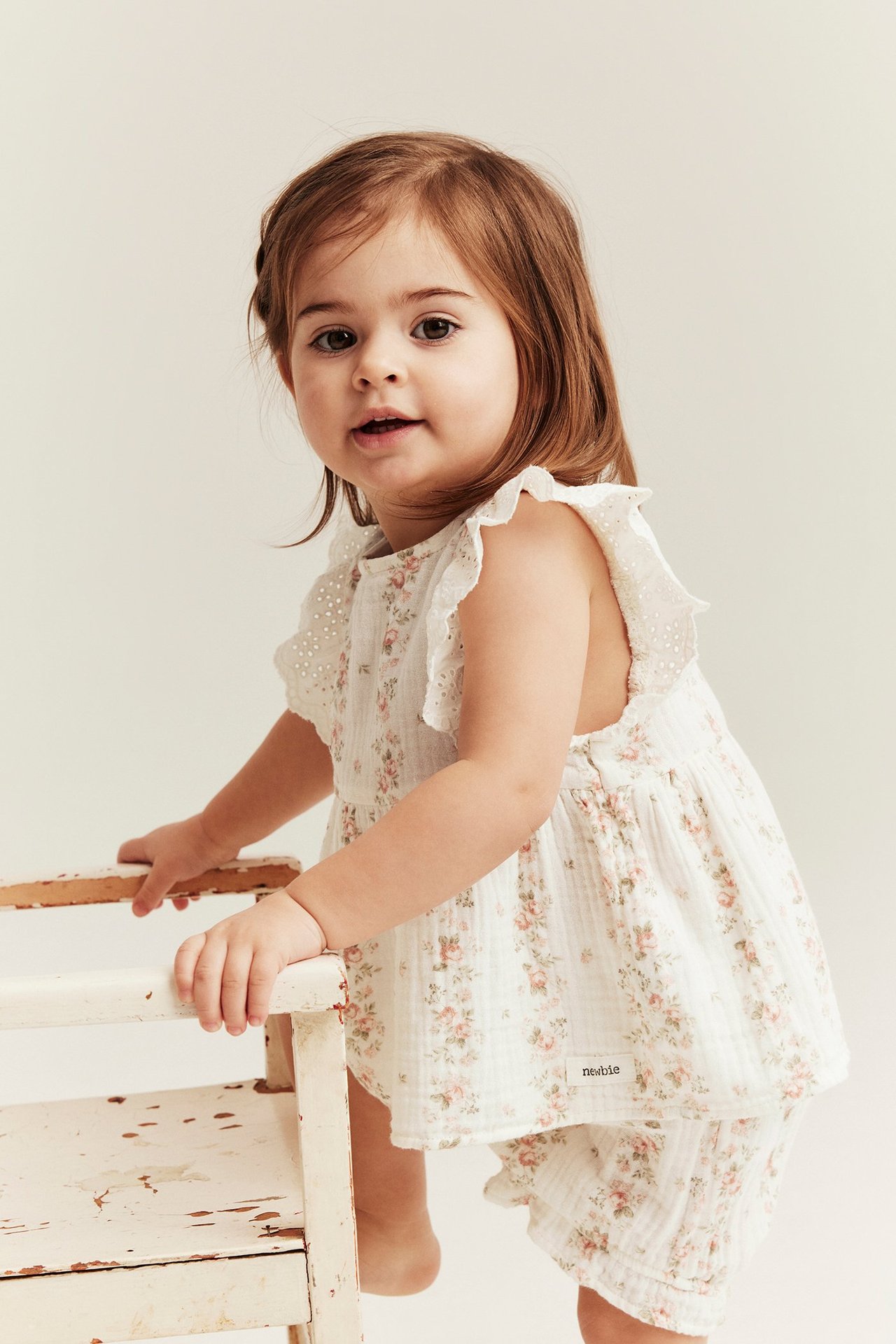 Vorderansicht: Ein Baby-Mädchen trägt ein cremeweißes Blumen-Musselin-Shorts-Set mit Peplum-Oberteil und Rüschenärmeln.