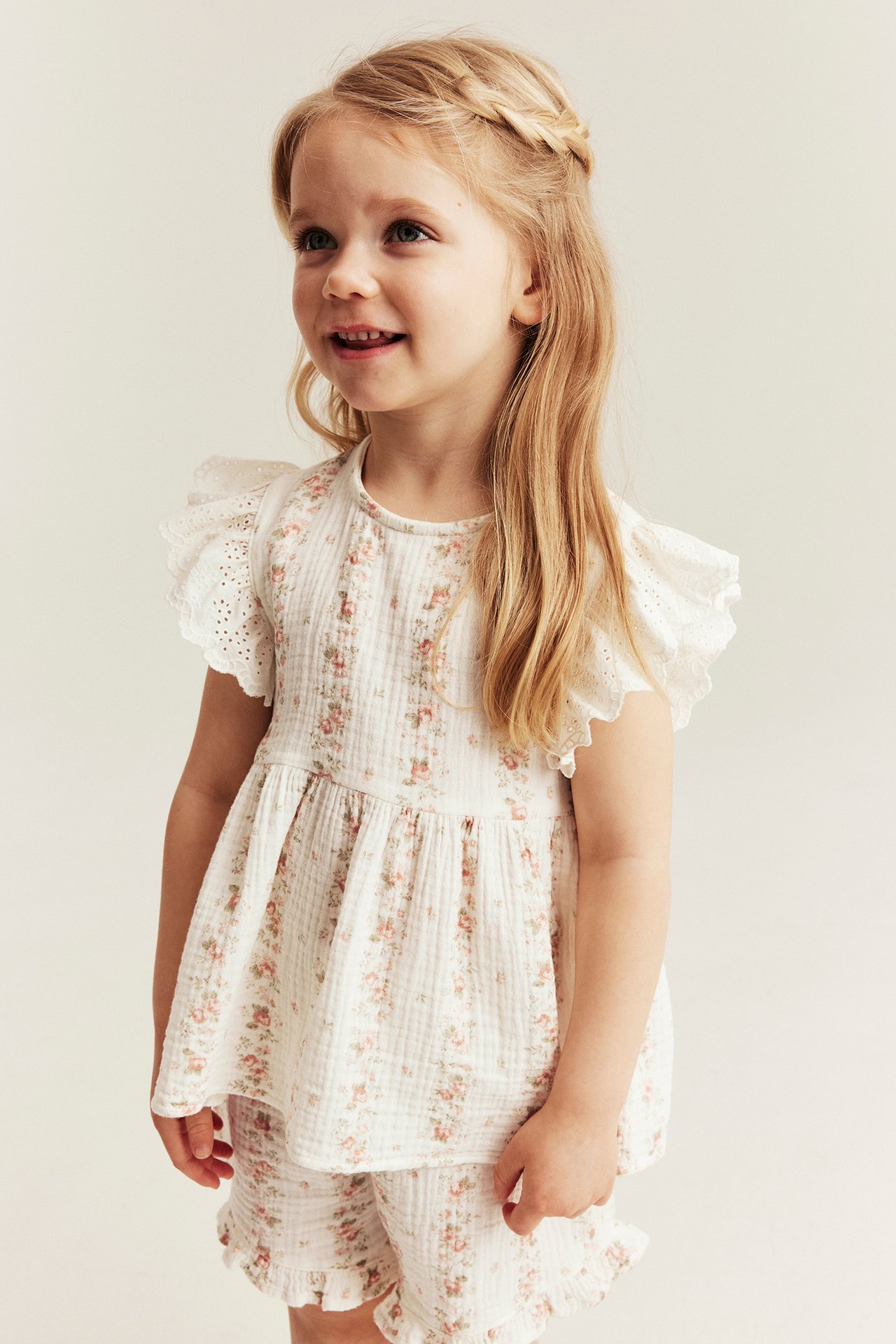 Front view: Kid girl in an off-white floral muslin peplum top with ruffle sleeves and matching shorts.