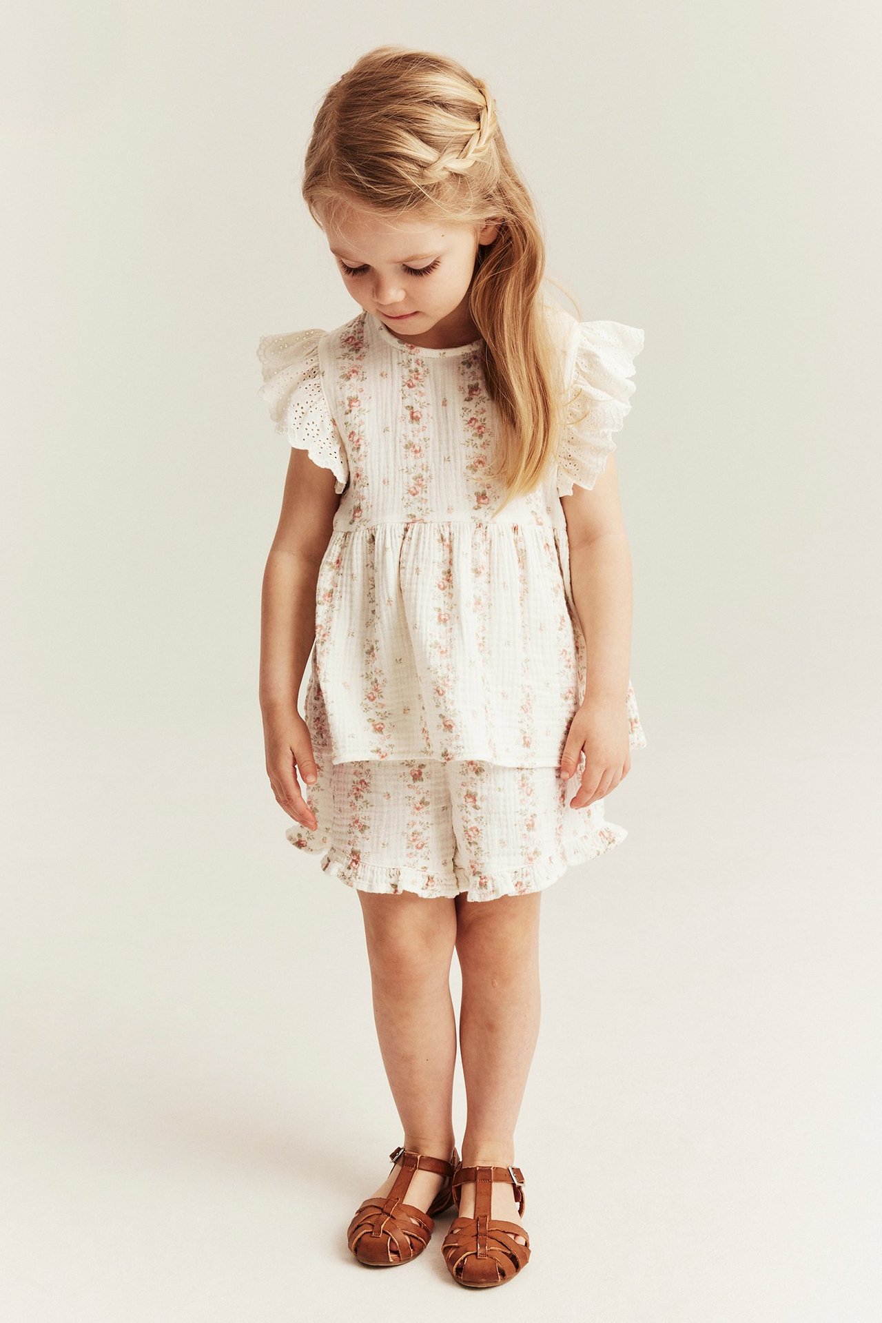 Front view: Kid wears an offwhite floral muslin top, matching ruffled shorts, and brown sandals.