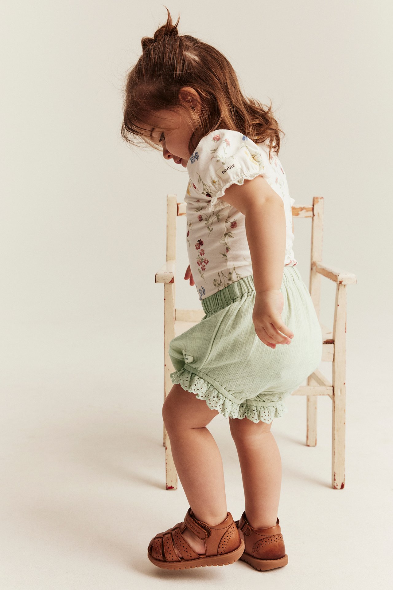 Side view of a kid in light green woven puff shorts with lace, a white floral top, and brown sandals.