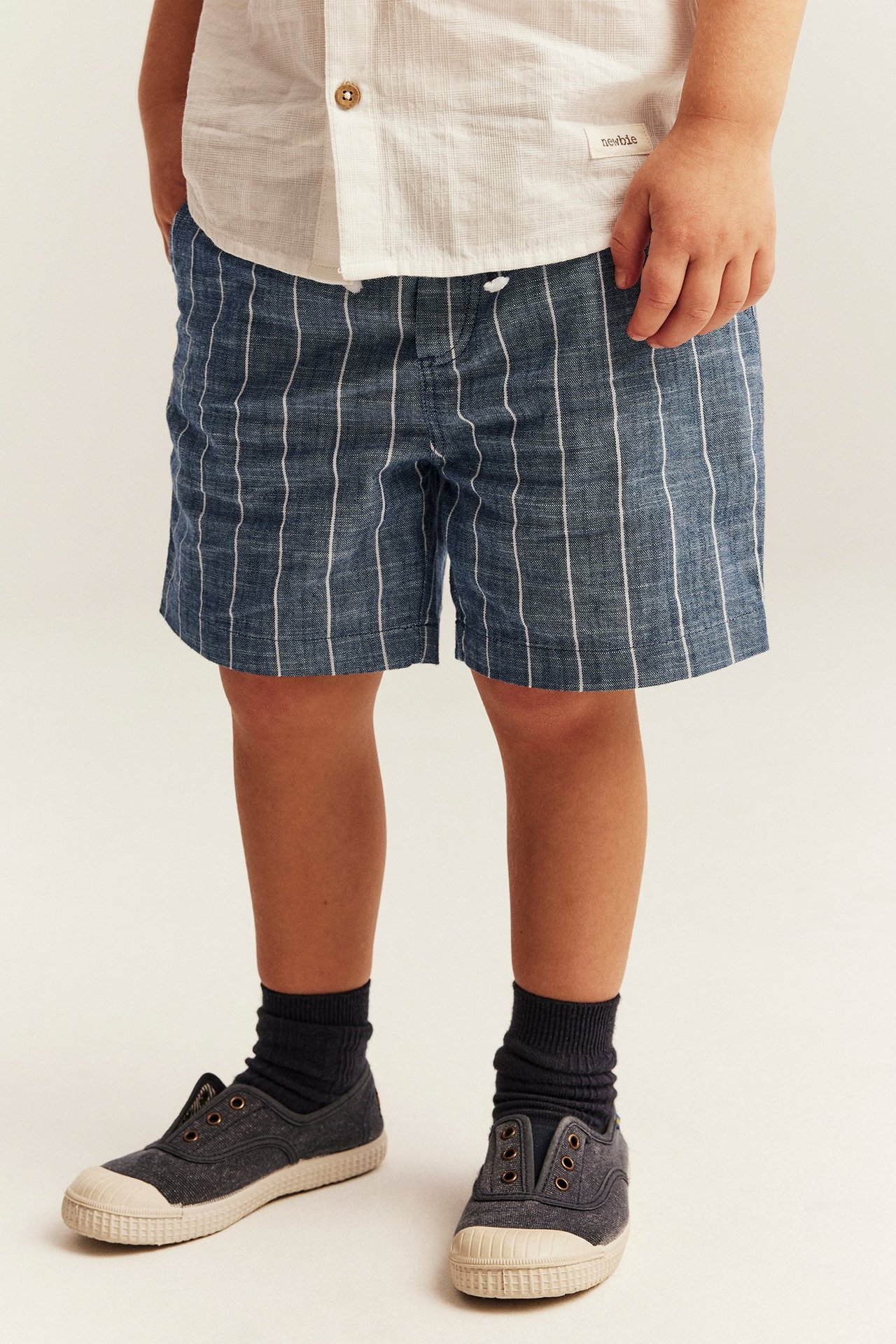 Blaue gestreifte Baumwollshorts mit Kordelzug, von Kind getragen mit weißem Hemd, dunklen Socken, Segeltuchschuhen.