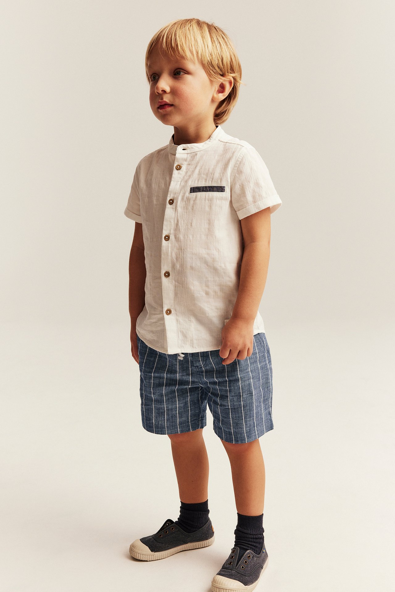 Front view of a kid wearing blue striped woven shorts, a white linen-blend shirt, dark socks, and grey canvas shoes.