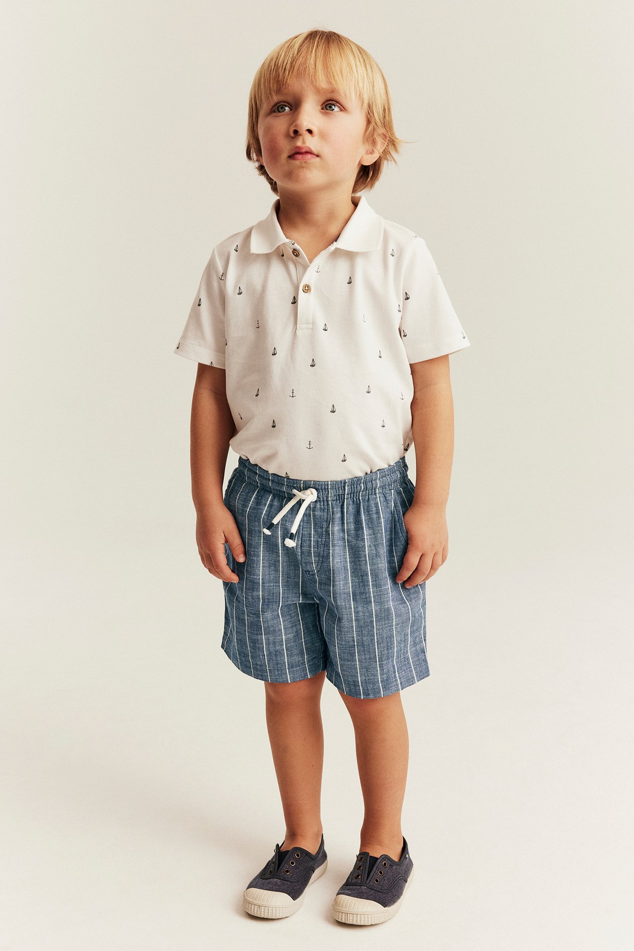 Front view of a kid wearing blue striped woven shorts with drawstring, white anchor polo, and dark blue shoes.