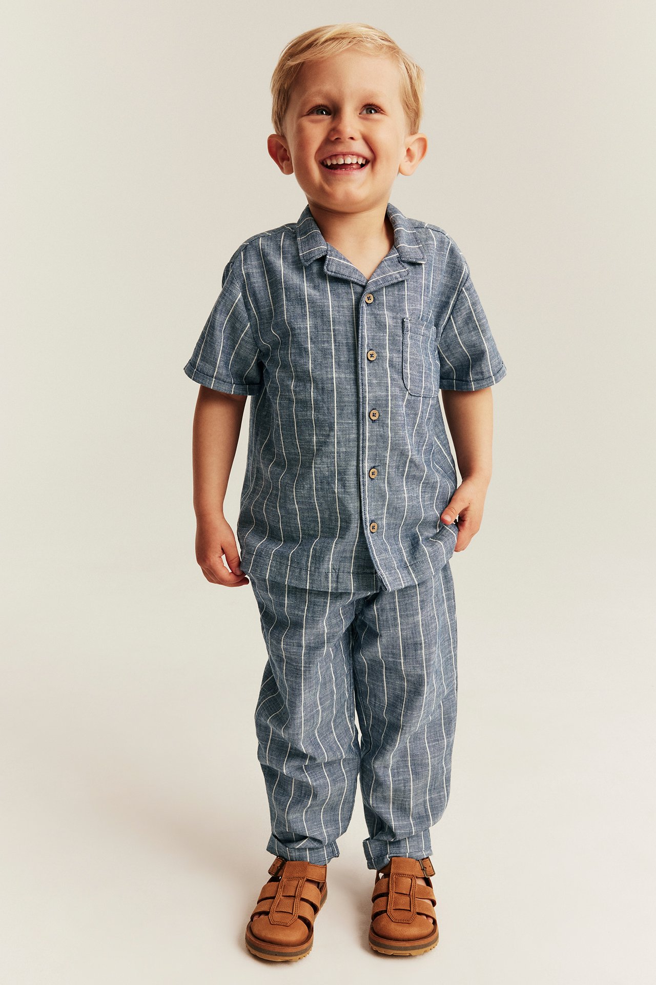 Front view of a smiling kid wearing a blue striped woven cotton shirt and matching trousers, with brown sandals.