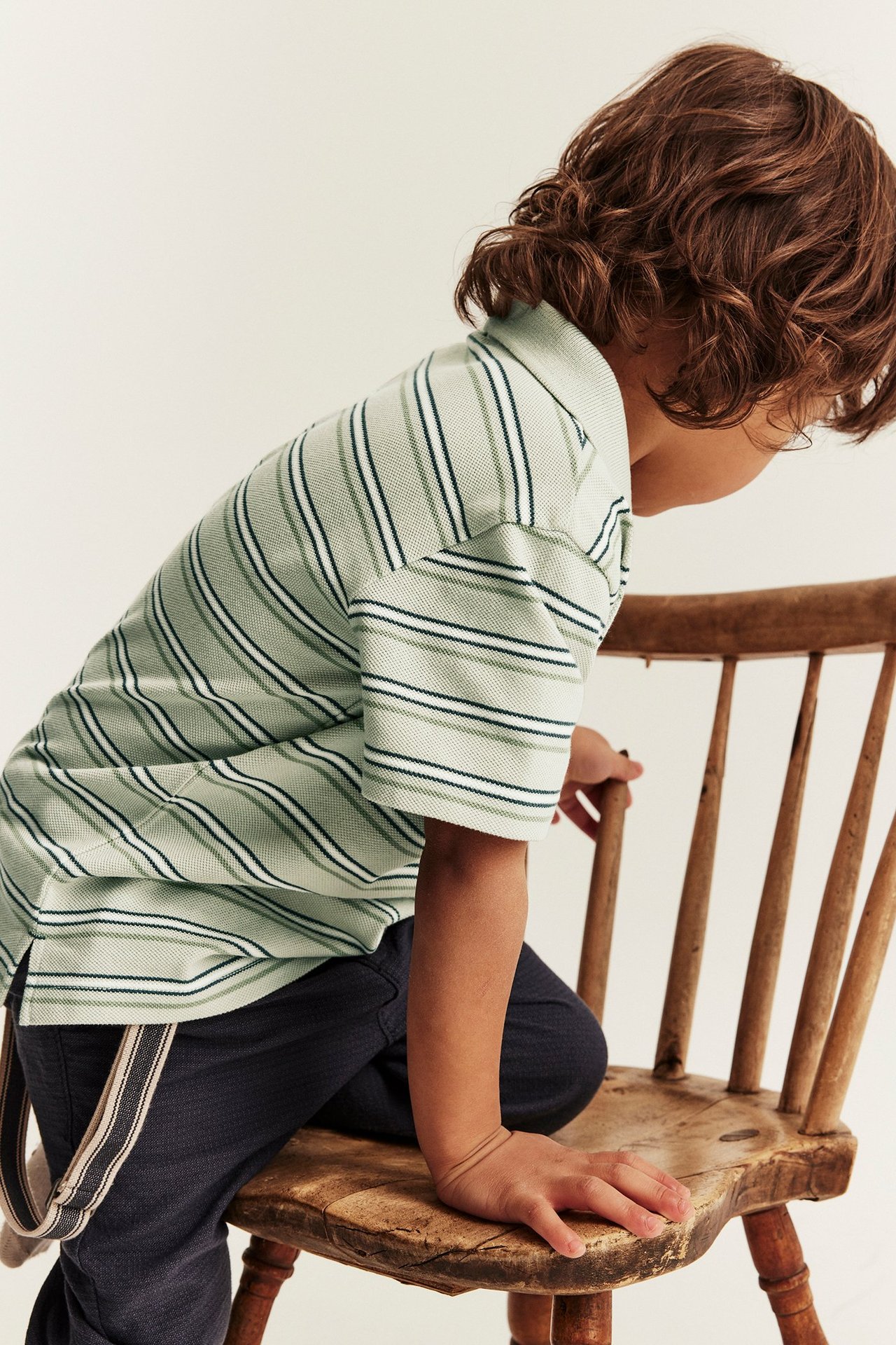Side view of a kid in a light green striped polo t-shirt and dark trousers with suspenders.