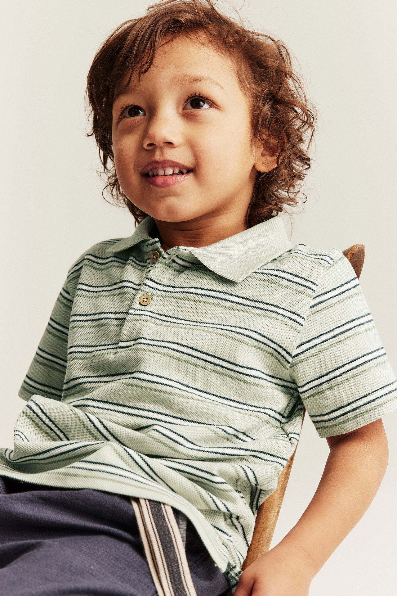Front view of a smiling kid in a light green striped cotton polo shirt and dark blue trousers.