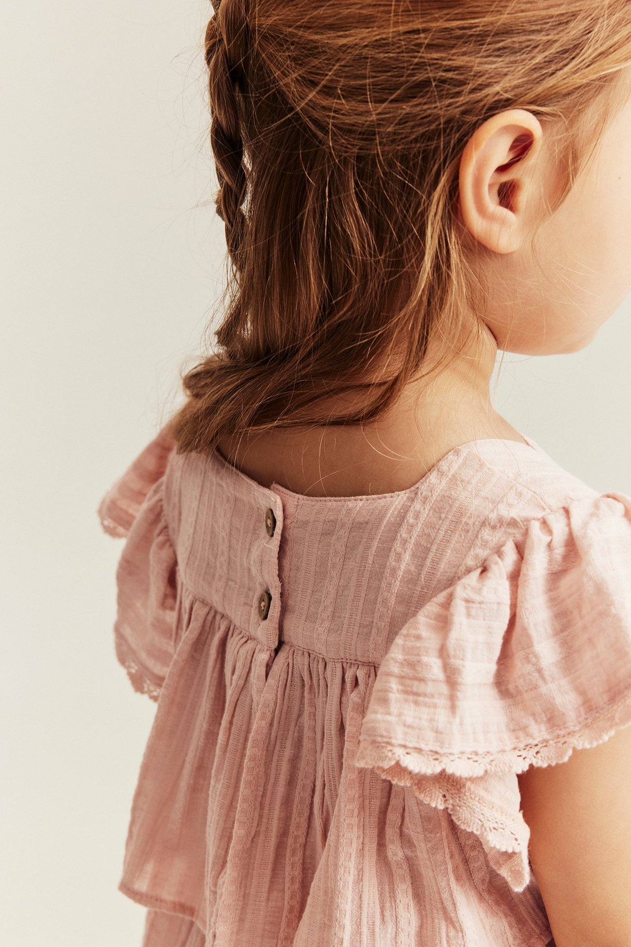 Back view of a kid in a light pink woven blouse with ruffled lace sleeves and back buttons.