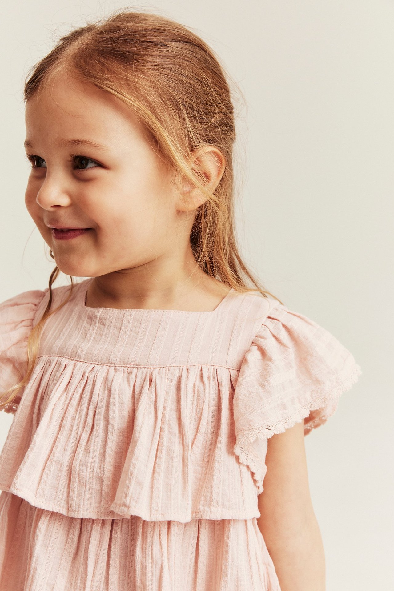 Close-up of a kid wearing a light pink ruffled woven blouse with a square neck and lace trim.