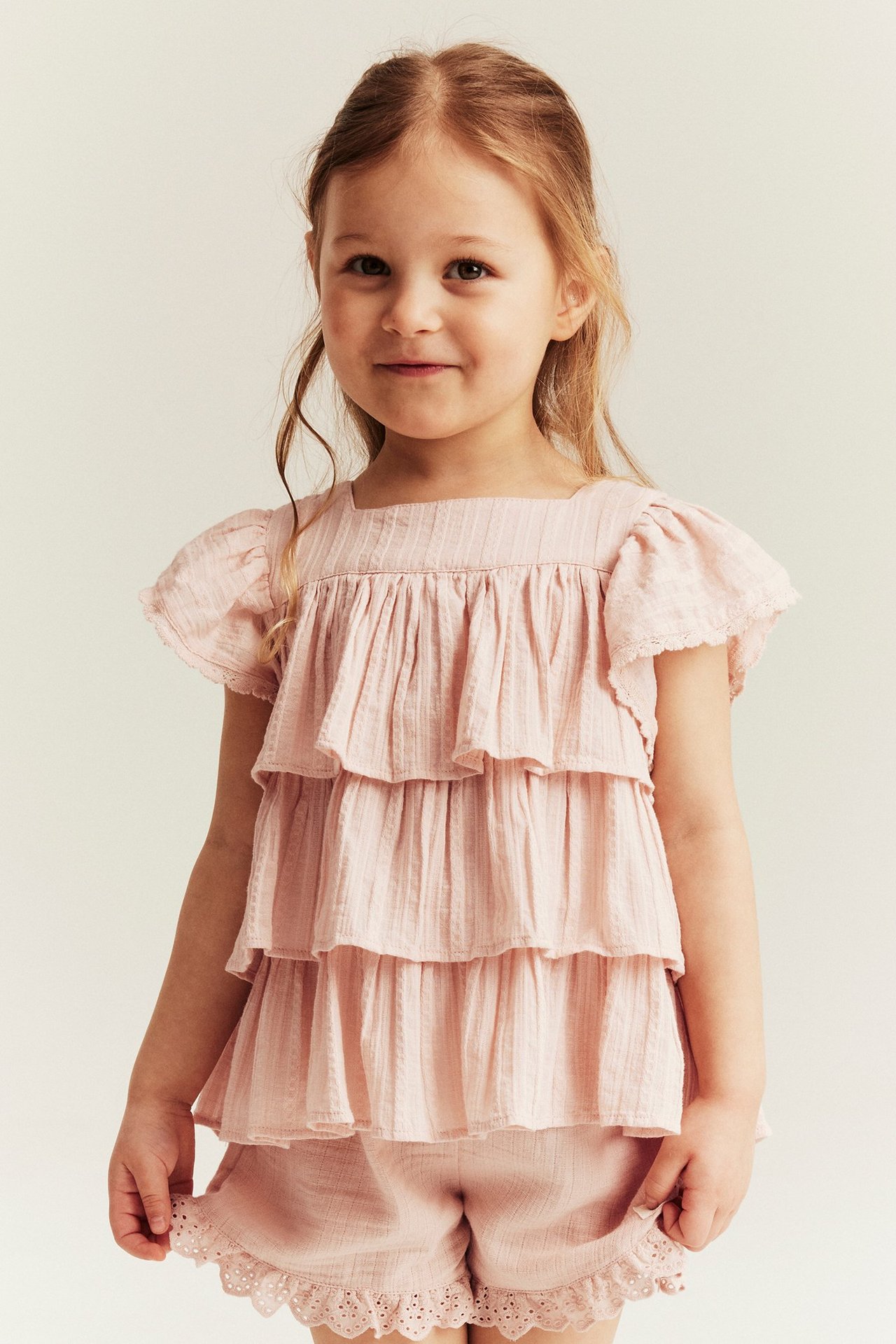 Front view of a kid in a light pink textured tiered ruffle blouse and matching eyelet shorts.