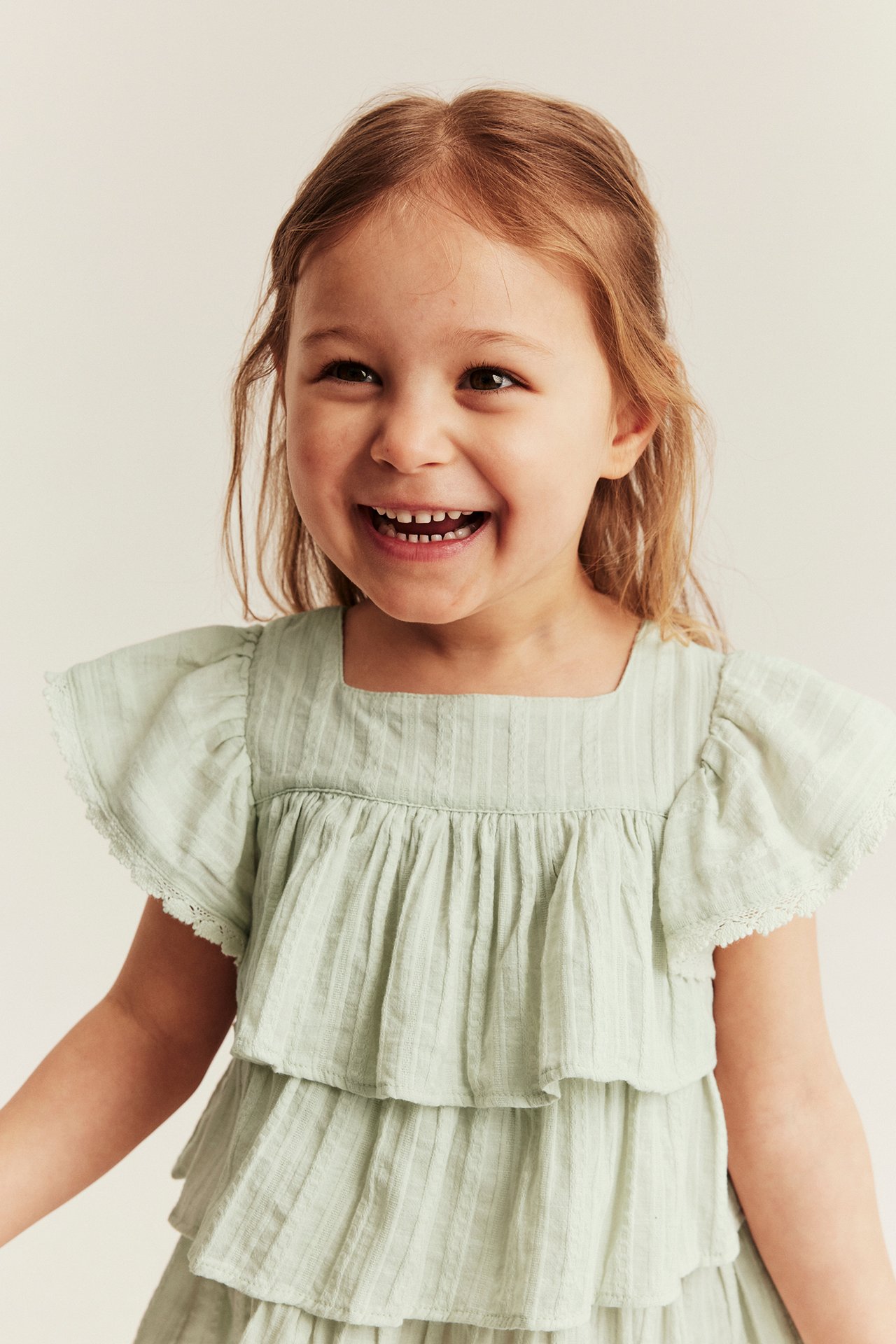 Front view: Kid in light green woven blouse with square neck, tiered ruffles, and lace sleeves.