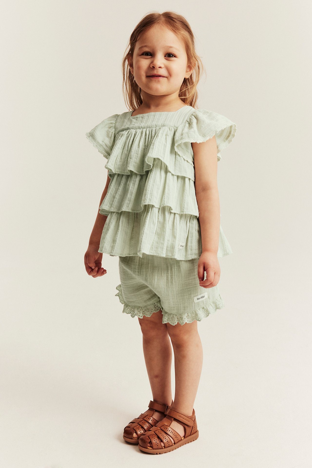 Front view: Kid in light green tiered ruffle blouse, matching shorts with lace, and brown sandals.