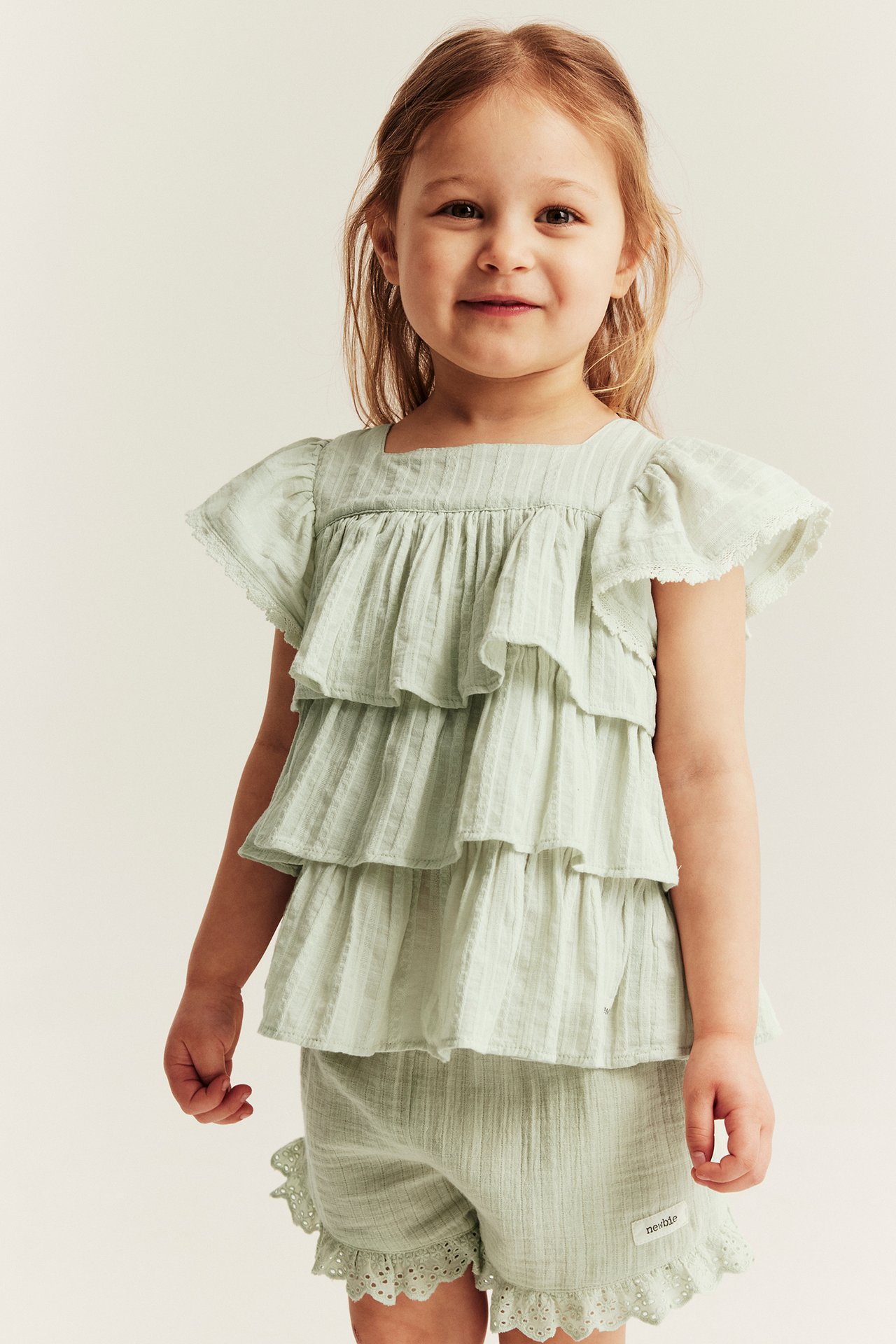 Front view of a kid wearing a light green tiered ruffle blouse with lace trim and matching textured shorts.