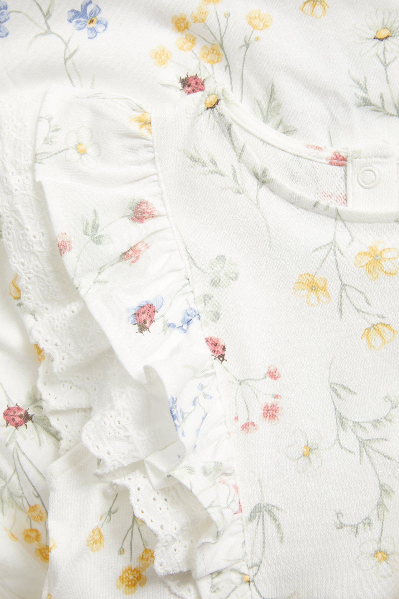 Close-up of an off-white floral jersey dress with ruffles, lace details, snap buttons, and ladybug print.