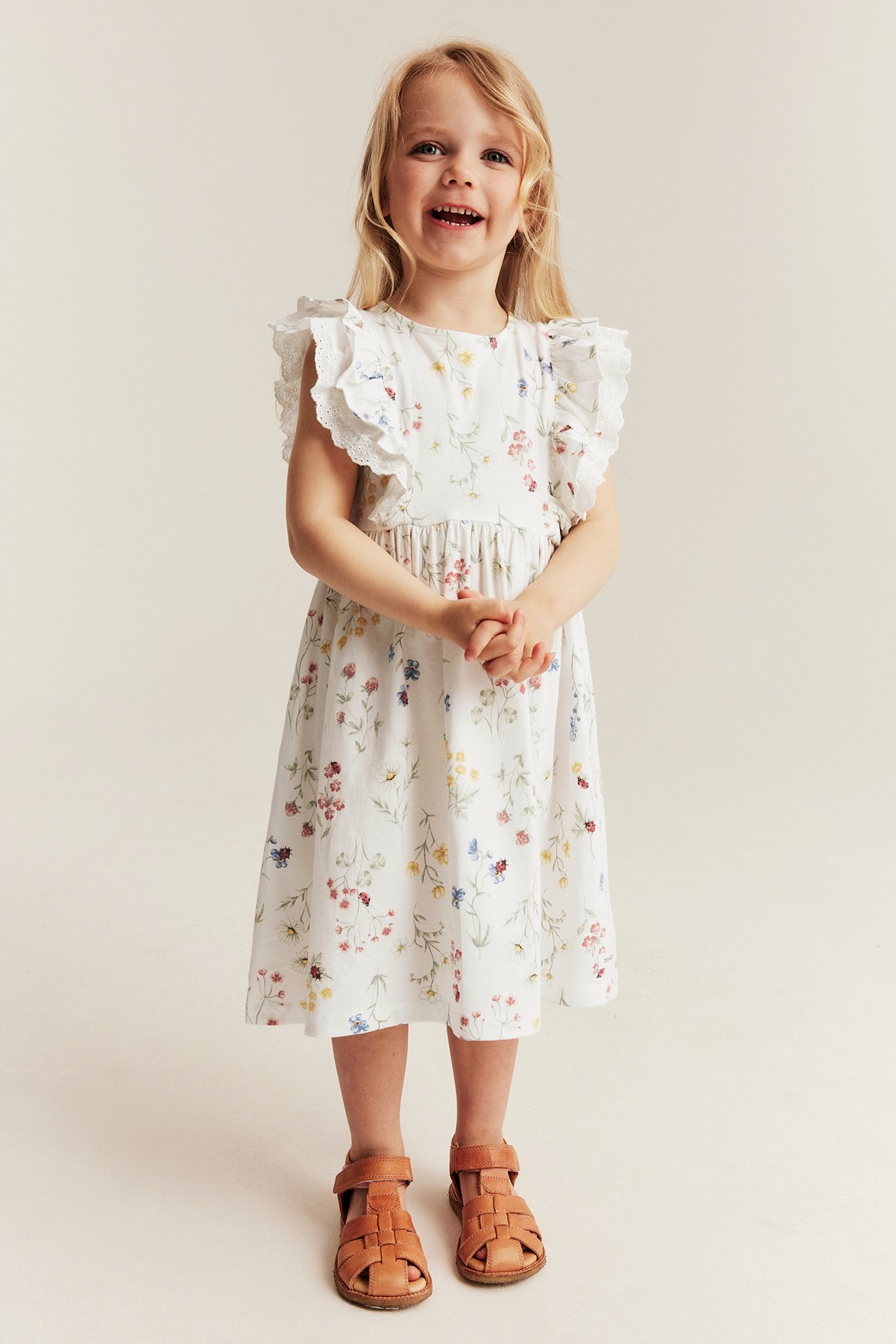 Frontansicht: Mädchen in offweißem Blumen-Jerseykleid mit Rüschenärmeln und hellbraunen Sandalen.