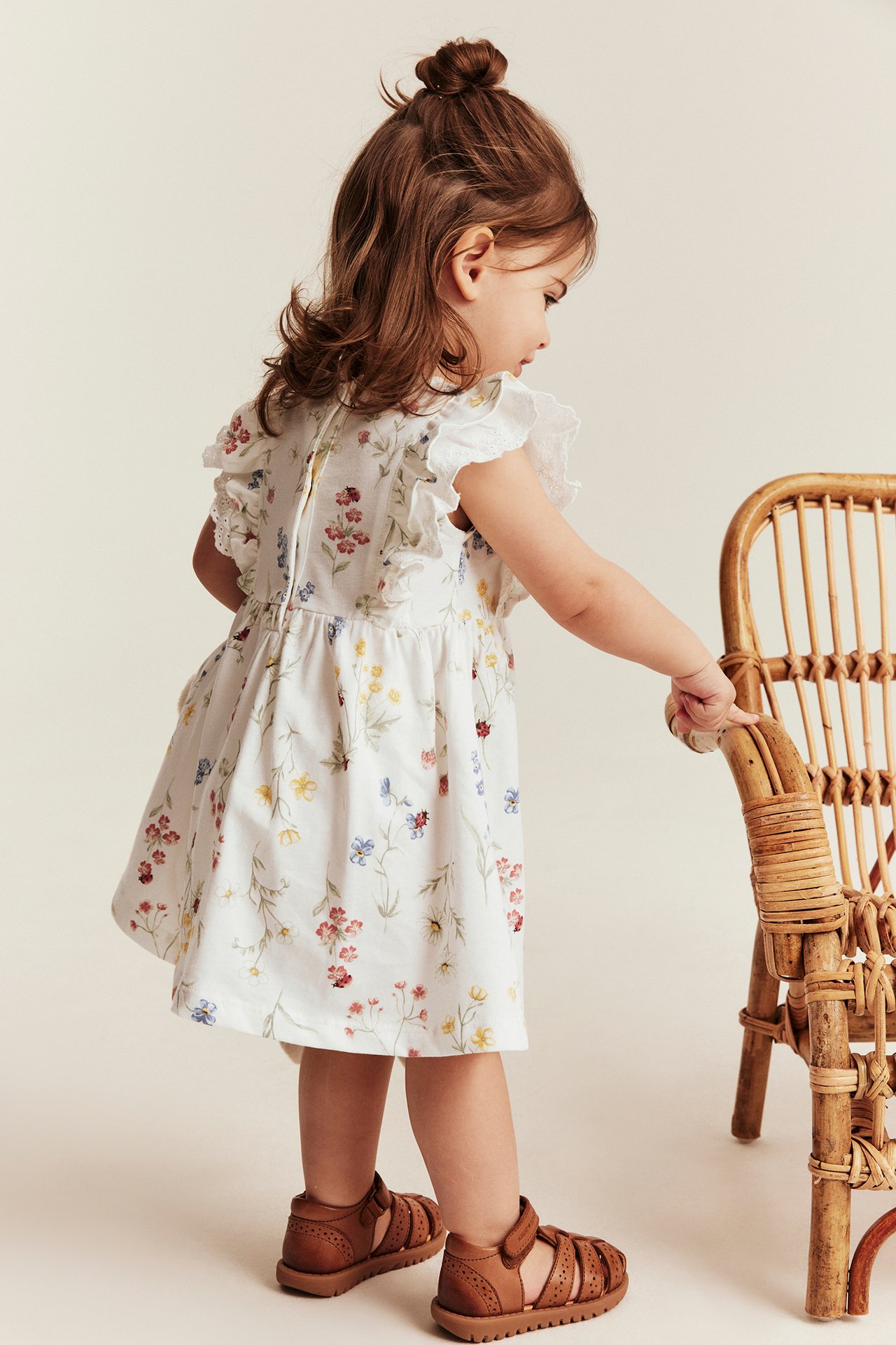 Side view: Kid in off-white floral jersey dress with ruffles, lace, snap buttons, brown sandals, touching chair.