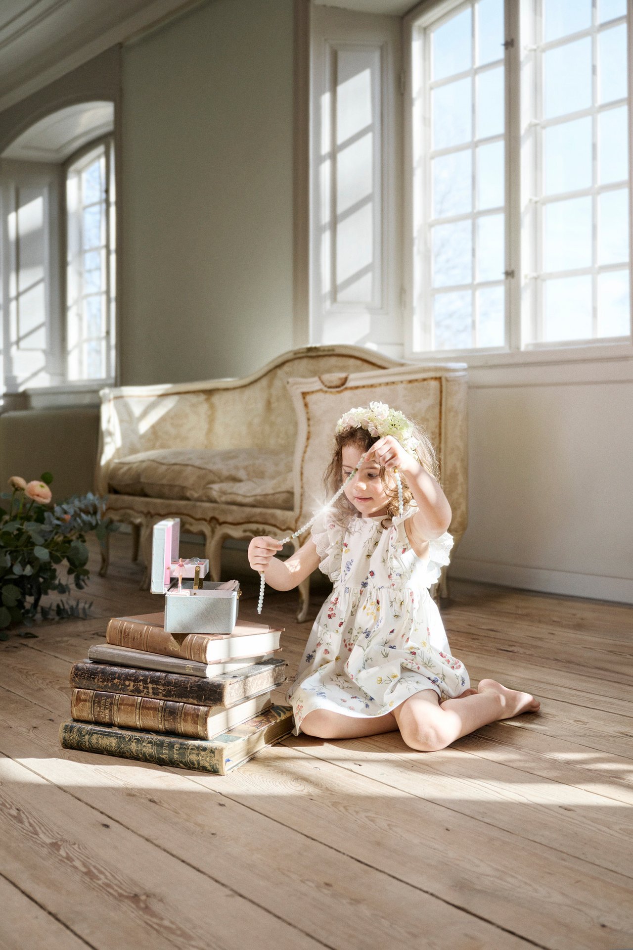 Front view: Kid girl in an off-white floral jersey dress with ruffles, wearing a flower wreath, holding pearls.