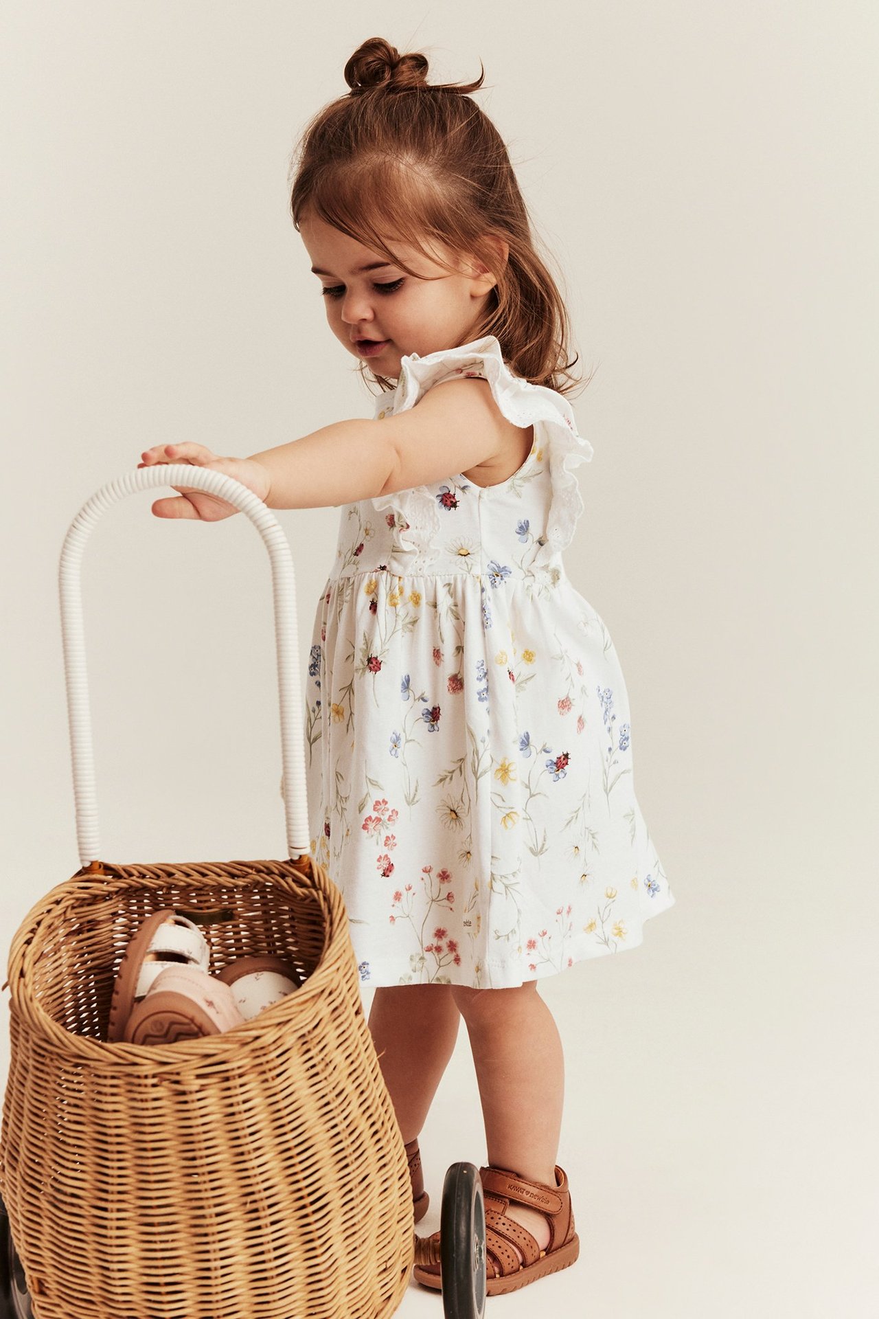 Seitenansicht Baby in cremefarbenem geblümten Jerseykleid, Rüschen, braune Sandalen, hält Korbtrolley.