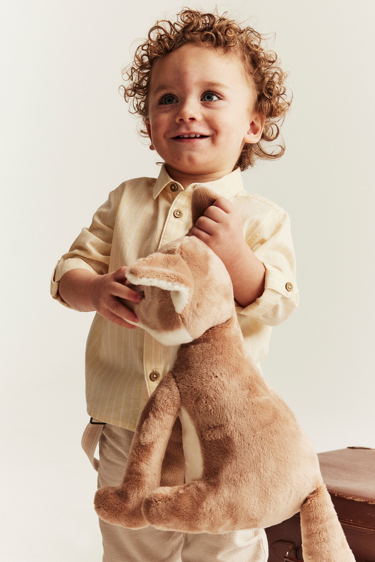 Front view: Kid in a yellow striped cotton woven shirt with rolled sleeves, light pants, holding a brown plush toy.