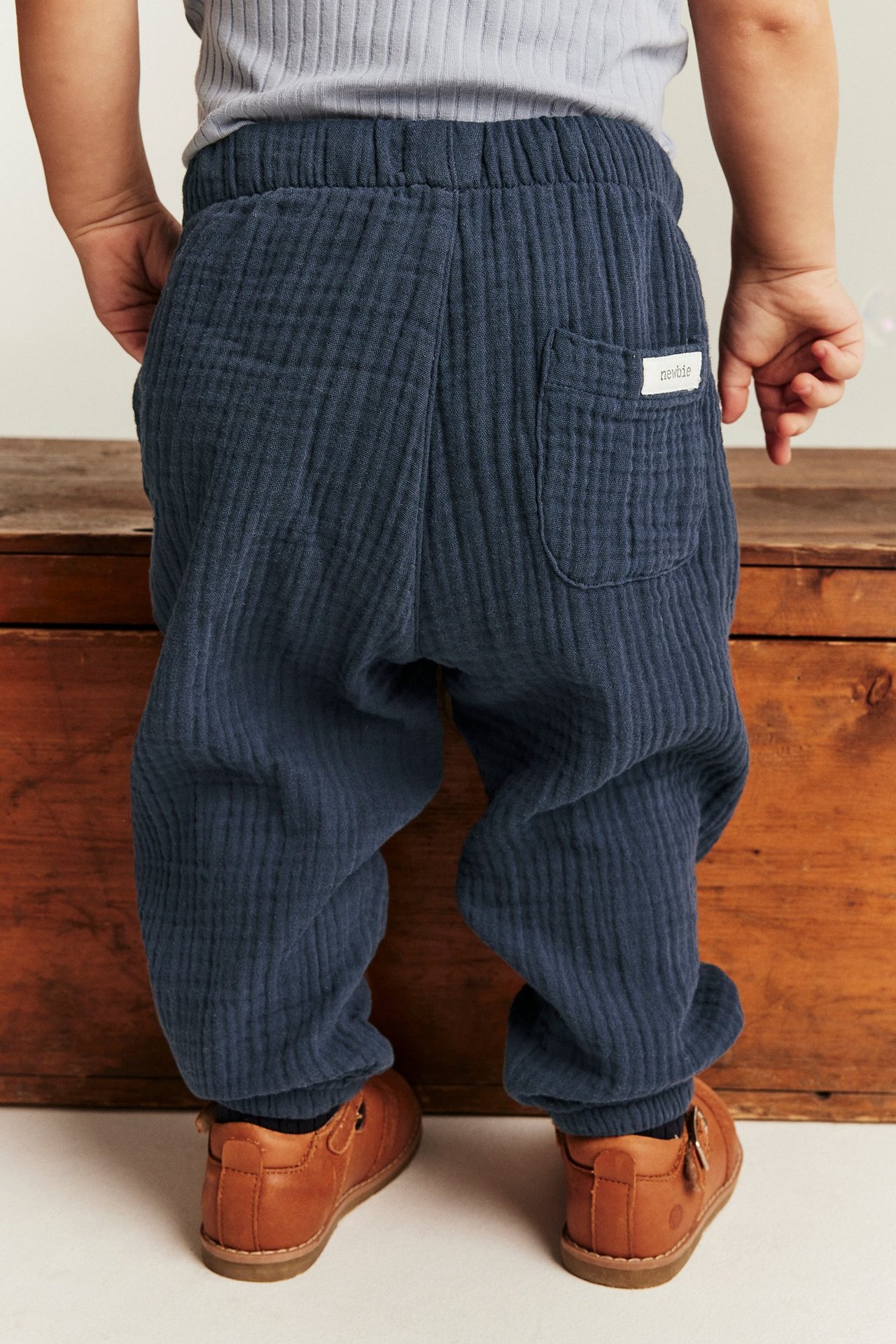 Rückenansicht eines Babys in dunkelblauen gewebten Musselinhosen mit Tasche, einem hellgrauen Oberteil und braunen Schuhen.