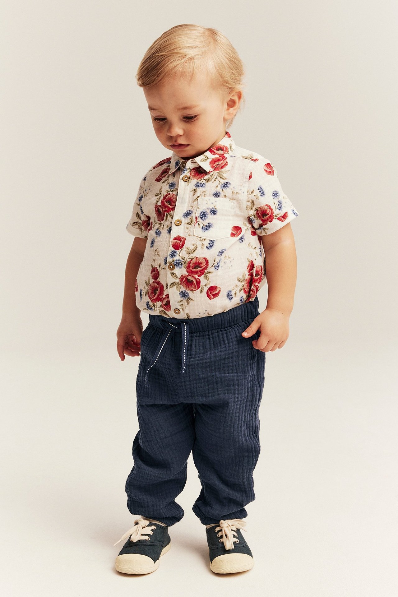 Vorderansicht: Babyjunge in dunkelblauer Musselin-Webhose, einem Blumenhemd und dunklen Turnschuhen.