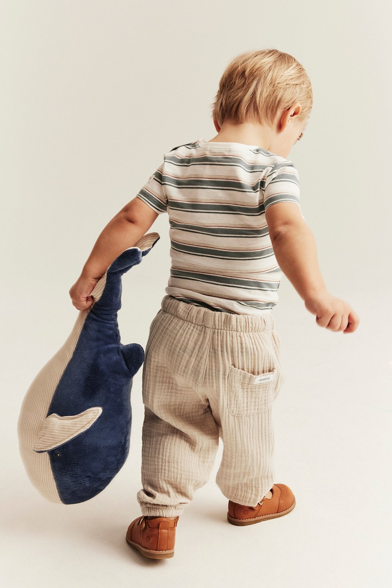 Rückenansicht eines Kindes in cremeweißen Musselin-Hosen, geringeltem T-Shirt, braunen Schuhen, hält blauen Wal-Plüsch.