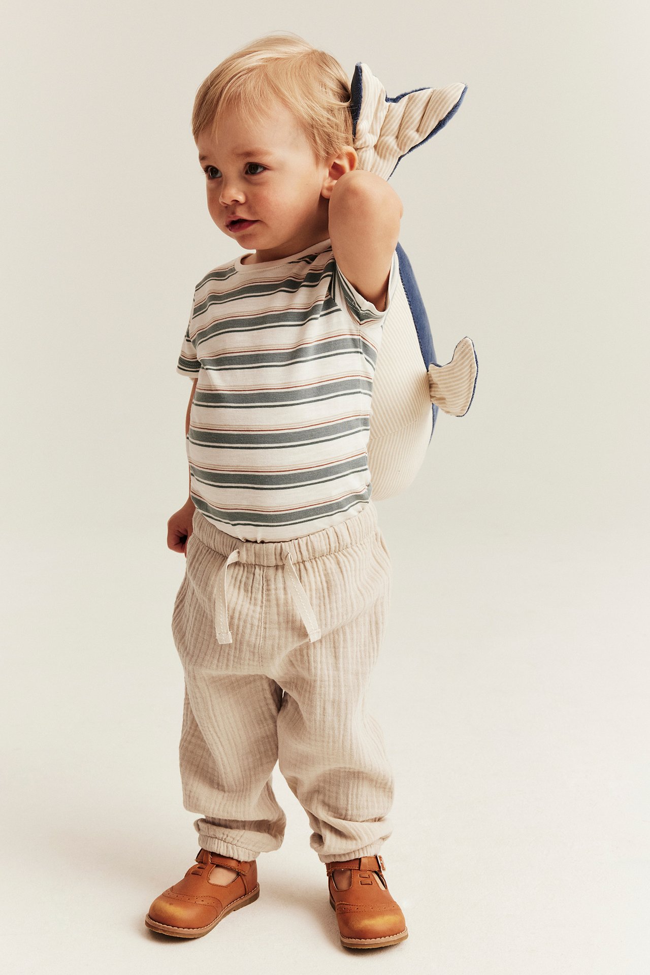 Vorderansicht eines Babys in cremeweißen Musselin-Hosen, gestreiftem Oberteil, braunen Schuhen und einem Plüschwal.