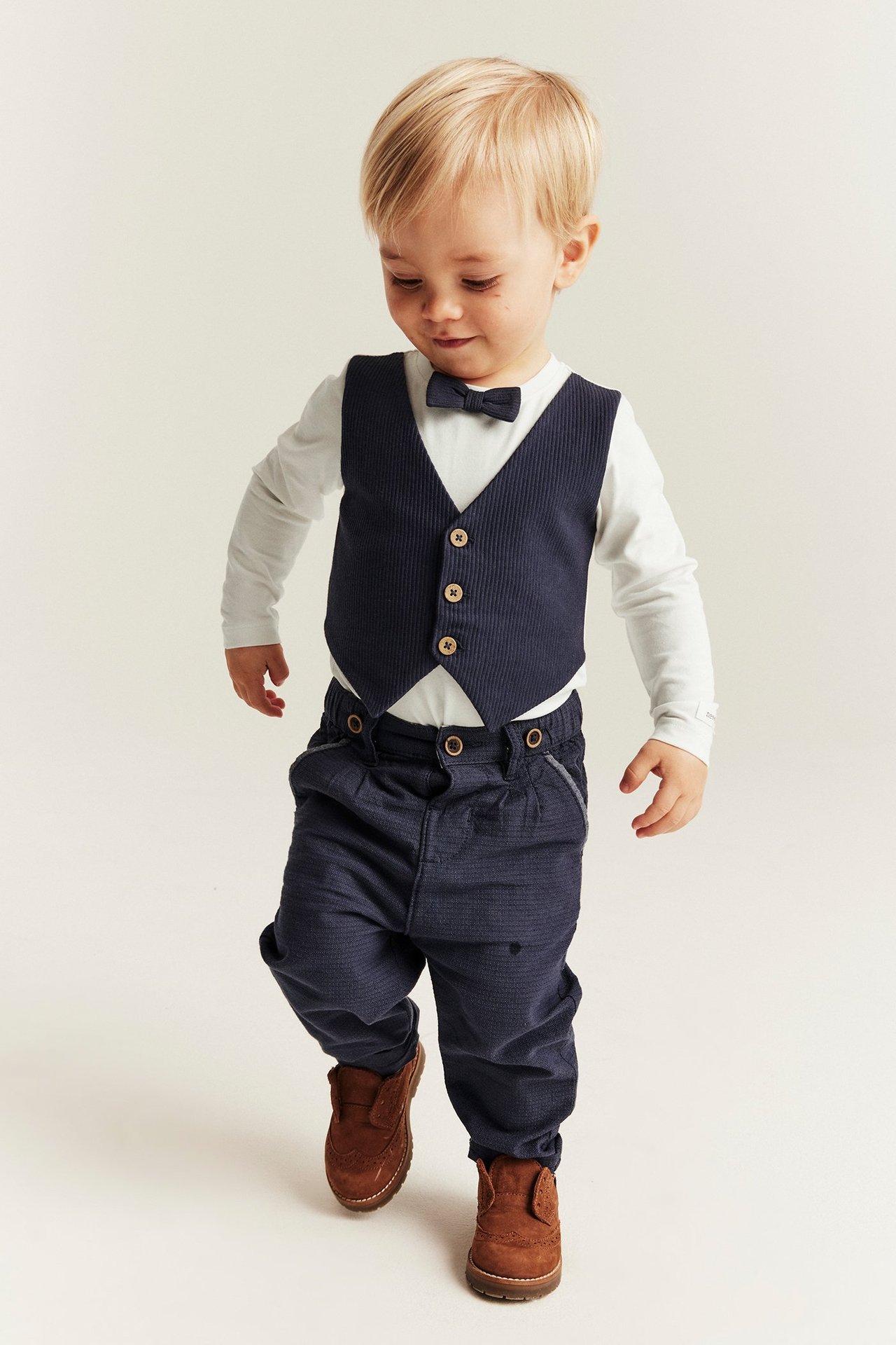 Front view: Baby boy in a white long-sleeve bodysuit, dark blue ribbed vest, bow tie, dark blue pants, brown shoes.