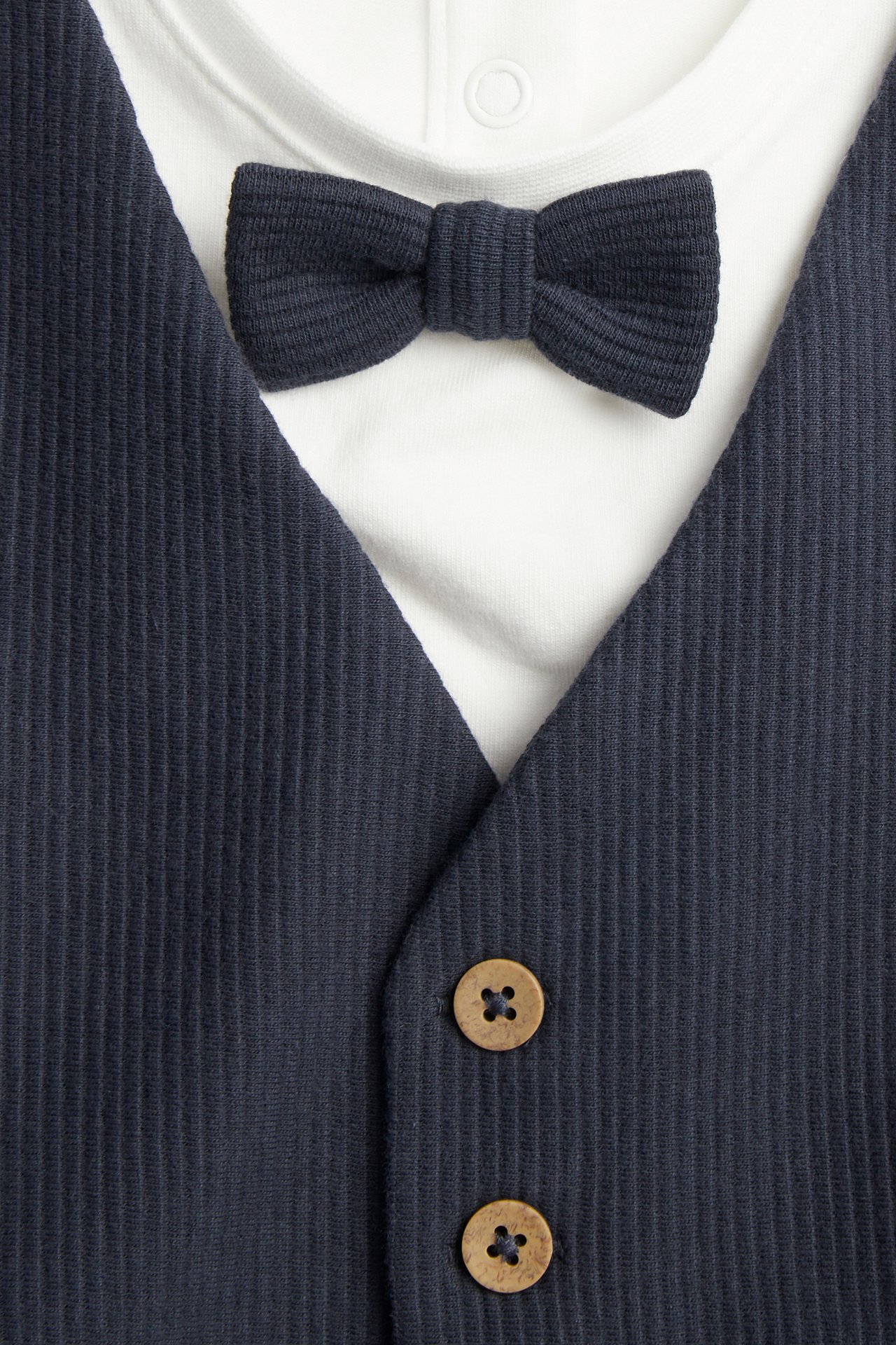 Close-up: baby's white bodysuit featuring attached dark blue ribbed vest, bow tie, and wooden buttons.