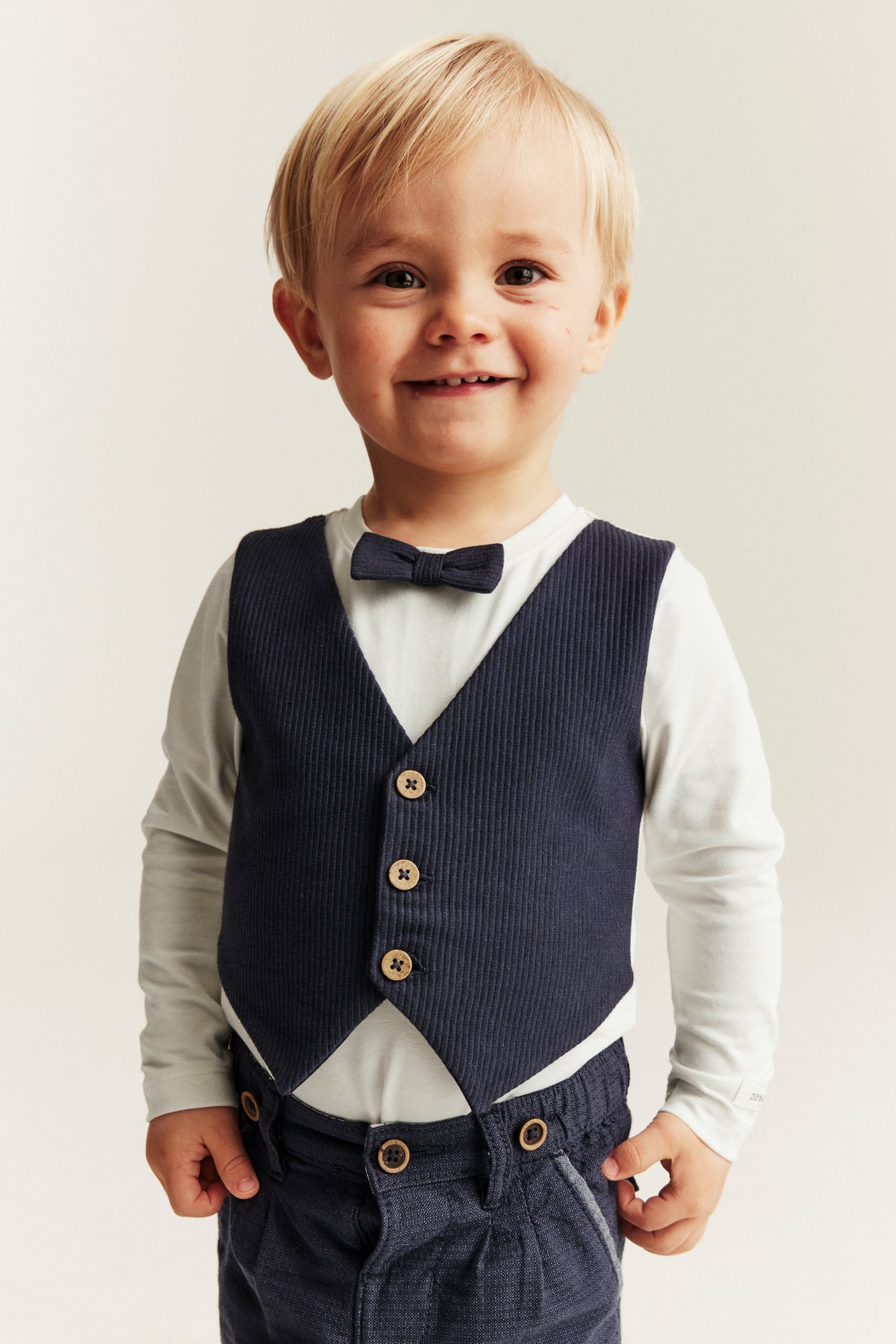 Front view of a baby boy wearing a dark blue ribbed vest bodysuit with light sleeves, bow tie, and dark blue trousers.