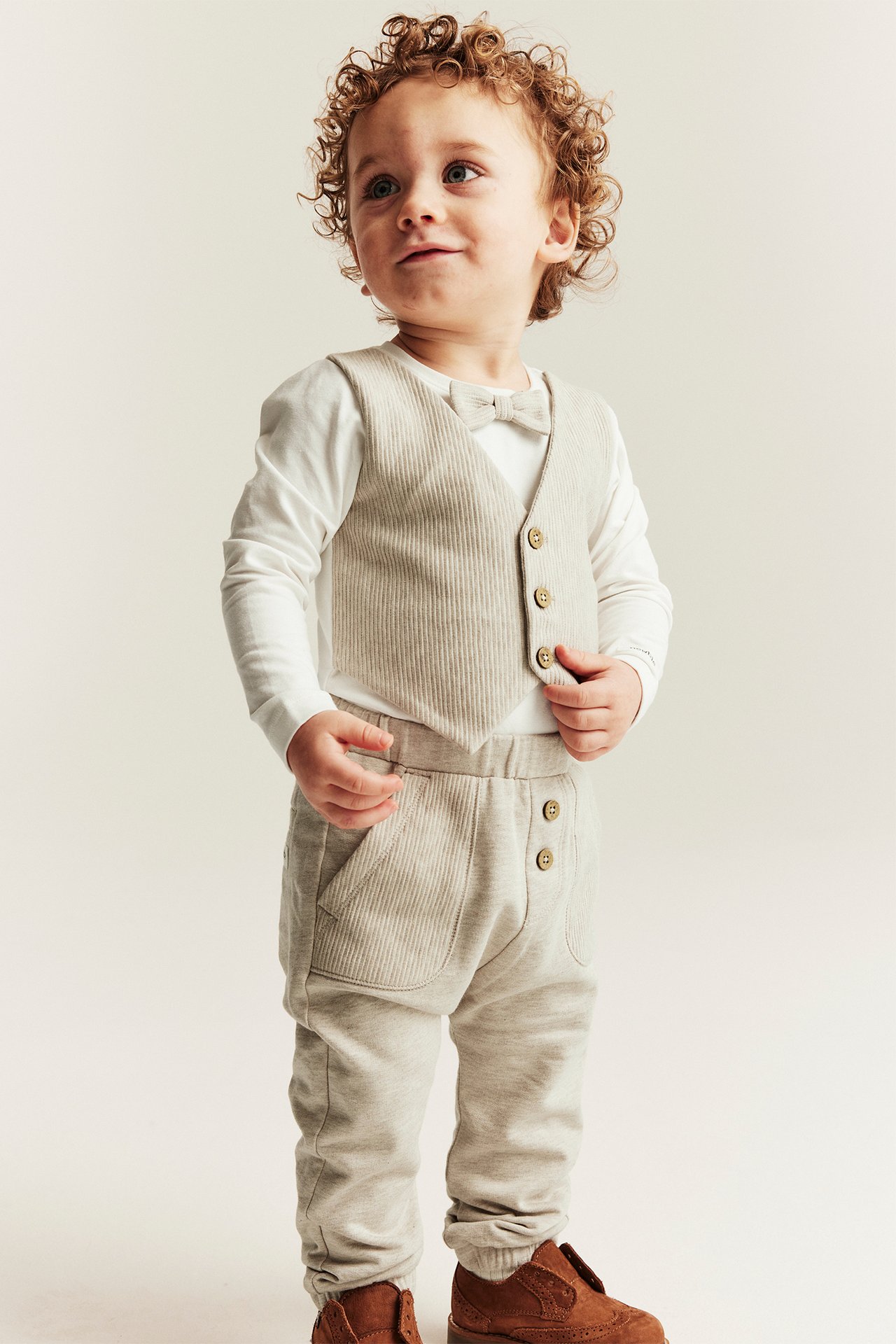 Front view: baby in beige melange ribbed bodysuit with attached vest, bow tie, white sleeves, grey trousers, brown shoes.