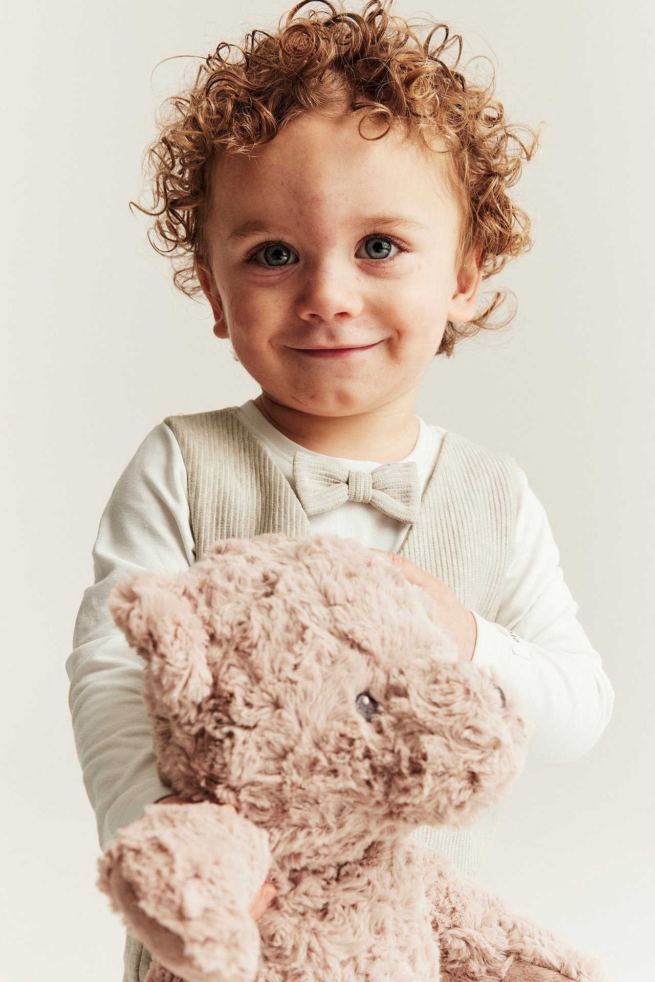 Vorderansicht: Lächelndes Baby trägt einen beigen Westenstrampler mit Fliege und hält einen weichen braunen Teddy.