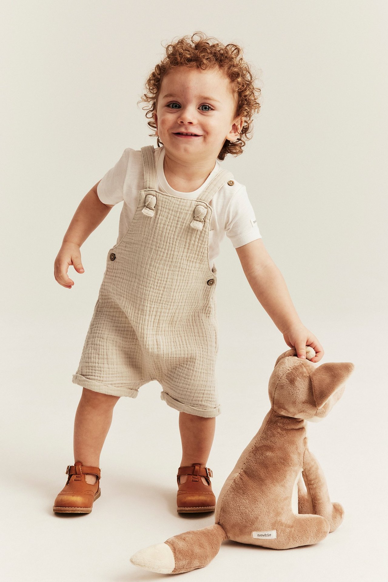 Vorderansicht: Baby trägt cremeweiße Musselin-Latzshorts, ein weißes T-Shirt und braune Schuhe, hält einen Plüschfuchs.