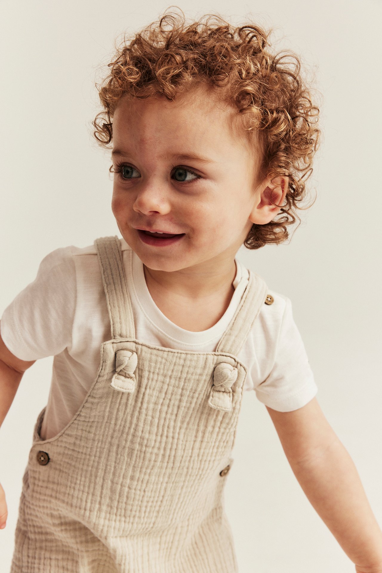 Close-up front view: baby in off-white woven muslin dungaree shorts with knot straps over a white t-shirt.