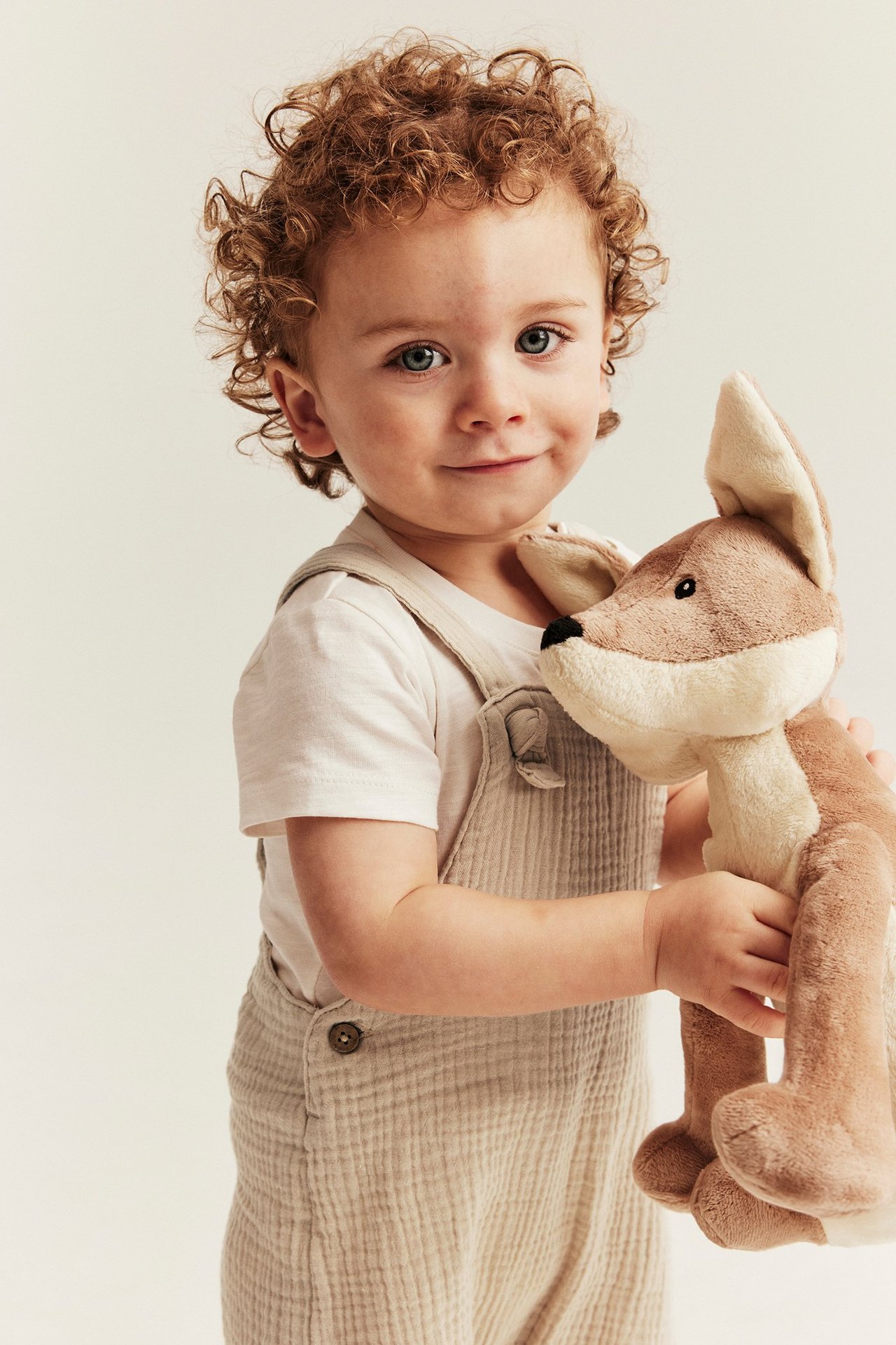 Vorderansicht eines Babys in cremefarbenen gewebten Musselin-Latzshorts über einem cremefarbenen T-Shirt, hält ein braunes Stofftier.
