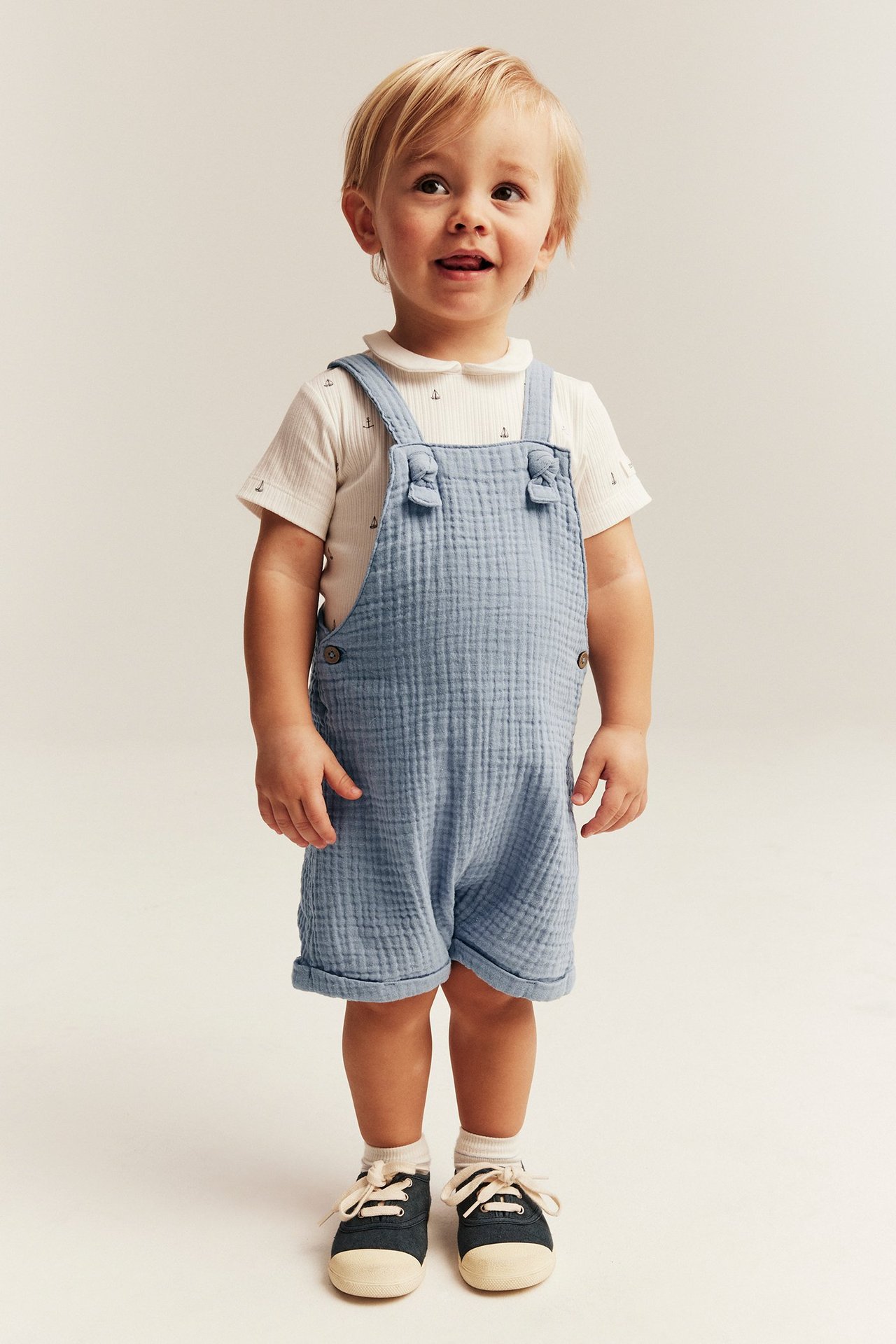 Front view: Baby in light blue muslin dungaree shorts with knots, white anchor tee, and dark shoes.