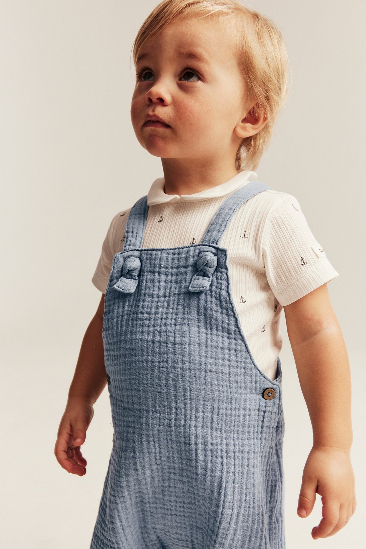 Front-side view of a baby in blue cotton muslin dungaree shorts over a white ribbed t-shirt with anchor pattern.