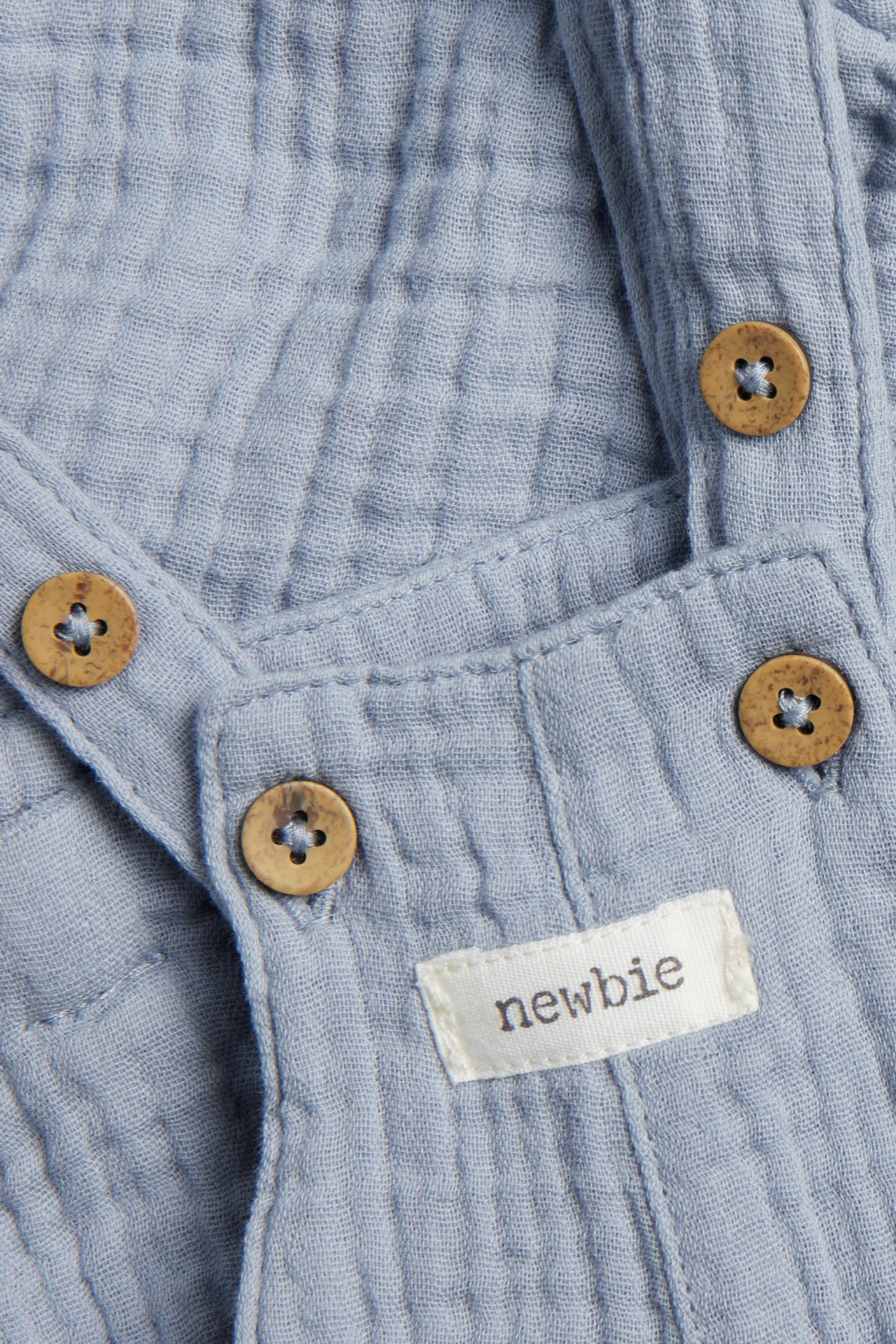 Nahaufnahme von hellblauen Baumwollmusselin-Latzshorts mit Holzknöpfen und Newbie-Etikett.
