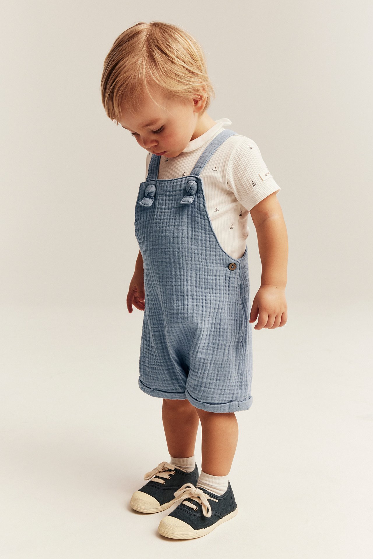 Vorderansicht: Baby in blauen Baumwoll-Musselin-Latzshorts, weißem Anker-Print-Top, dunkelblauen Schuhen, weißen Socken.