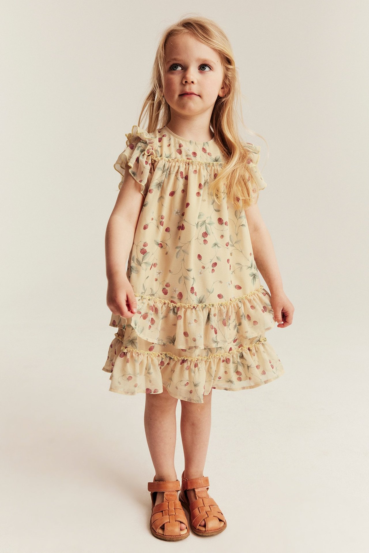 Vorderansicht: Mädchen trägt ein hellgelbes Erdbeermuster-Rüschenkleid aus Chiffon und braune Sandalen.