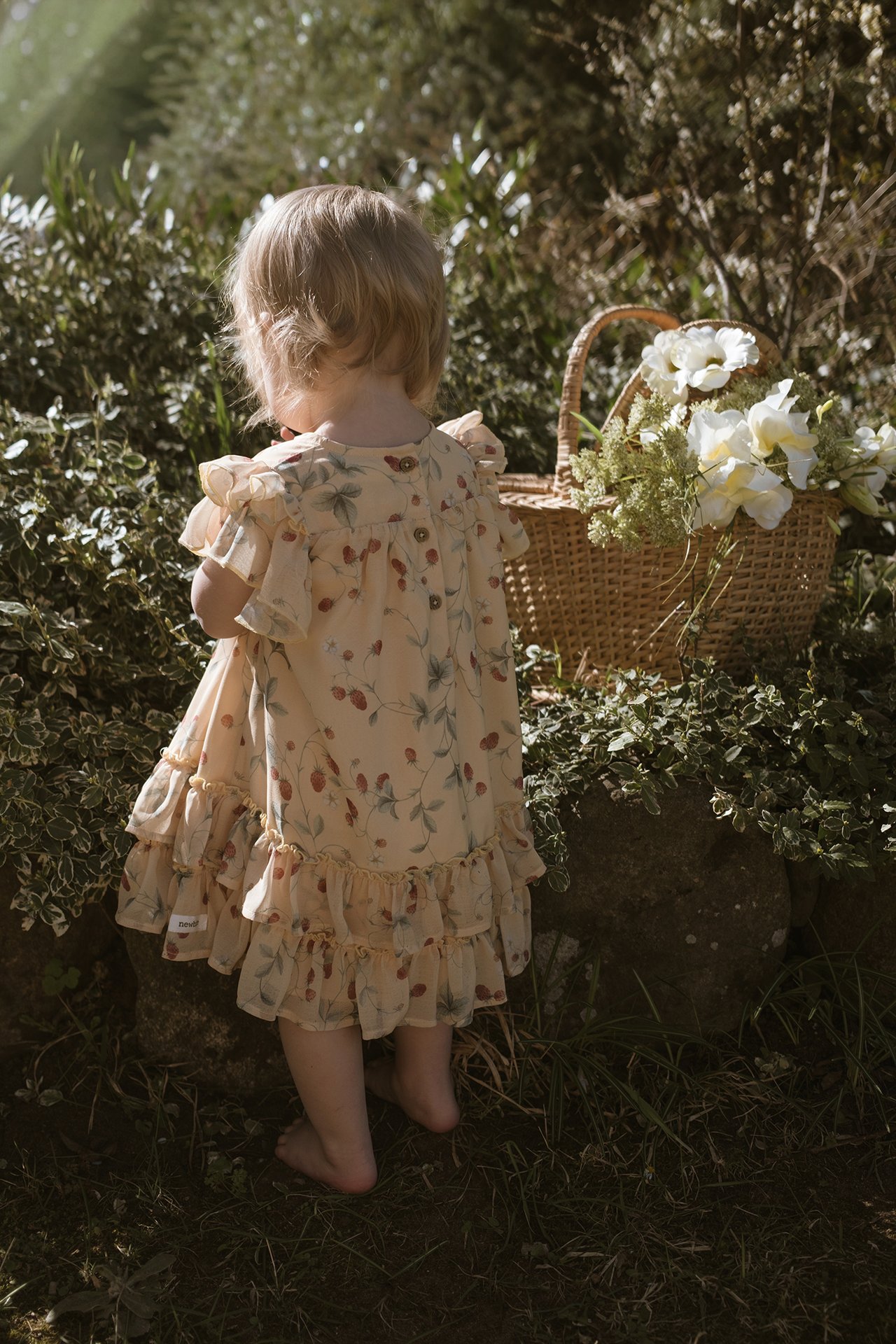 Back view of a baby in a yellow strawberry-patterned chiffon dress with ruffles and back buttons.