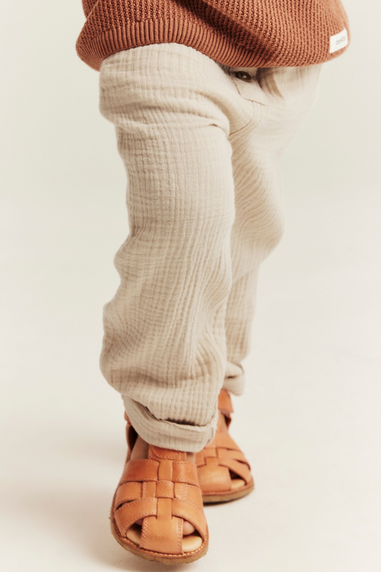 Vorderansicht: Baby trägt cremeweiße Baumwoll-Musselin-Hose mit hochgekrempelten Säumen, braunes Strickoberteil und braune Sandalen.