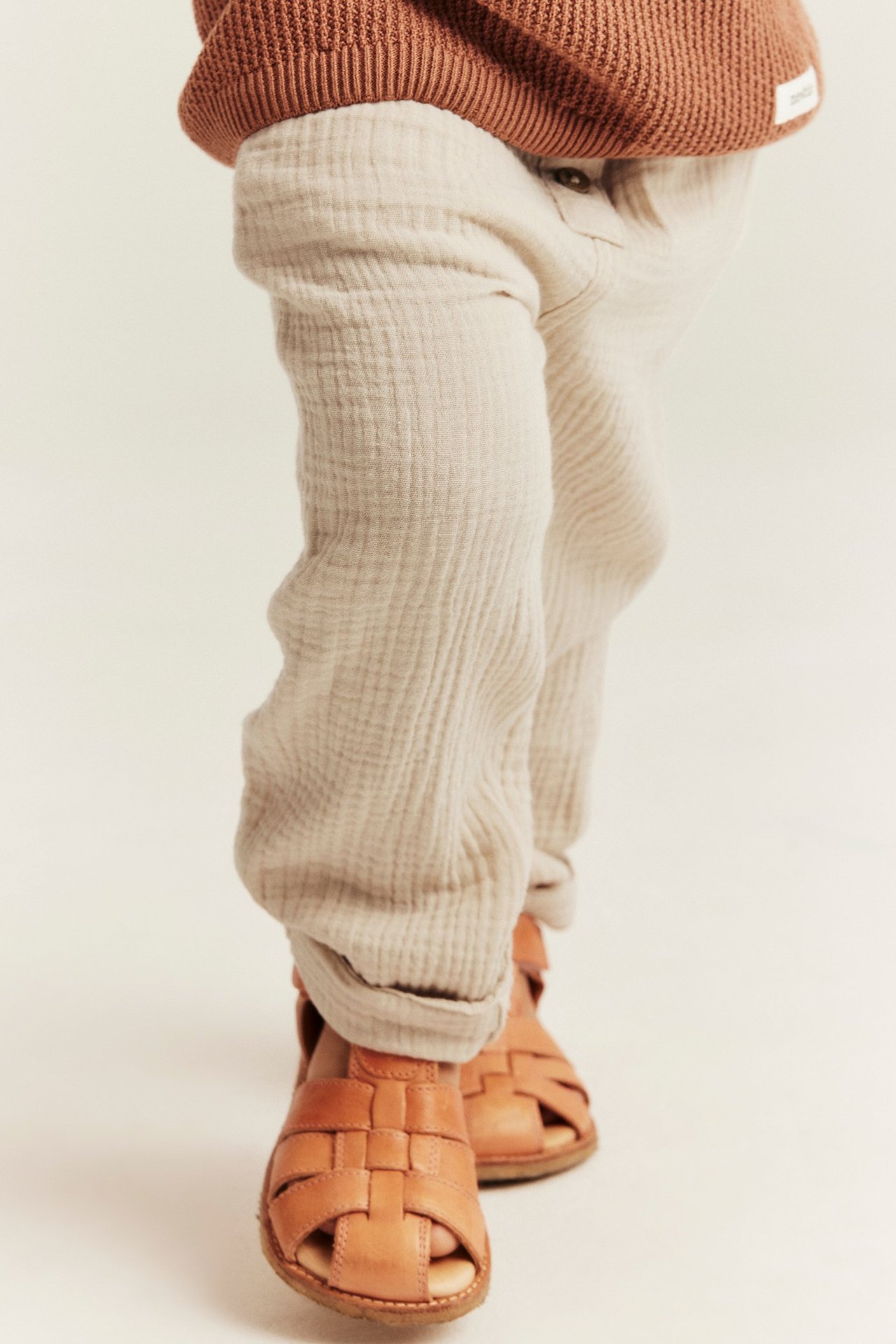 Vorderansicht: Baby trägt cremeweiße Baumwoll-Musselin-Hose mit hochgekrempelten Säumen, braunes Strickoberteil und braune Sandalen.