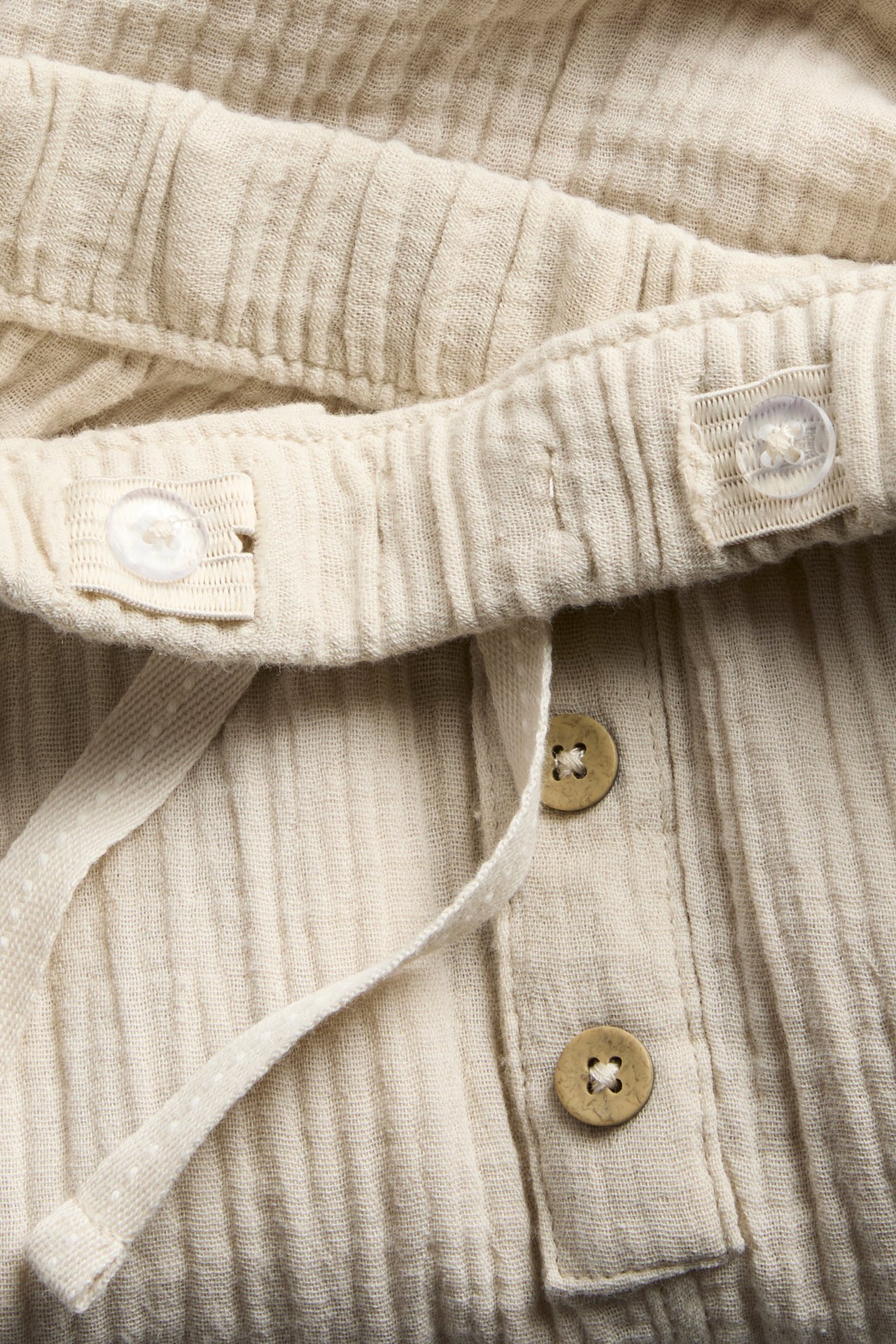 Nahaufnahme von cremeweißer Baumwoll-Musselin-Hose mit verstellbarem Gummibund, dekorativem Kordelzug und Knöpfen vorne.